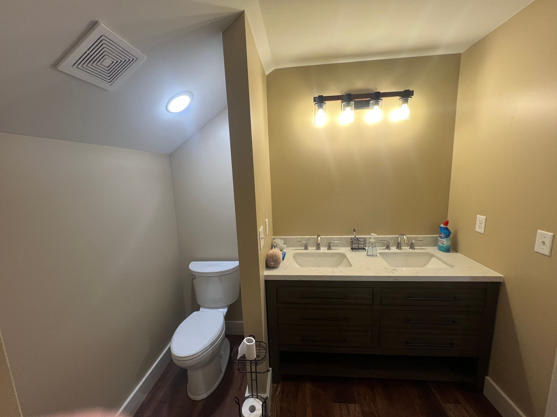 Bathroom with a toilet and double sink vanity, tan walls, dark wood cabinets, and a dark-colored light fixture.