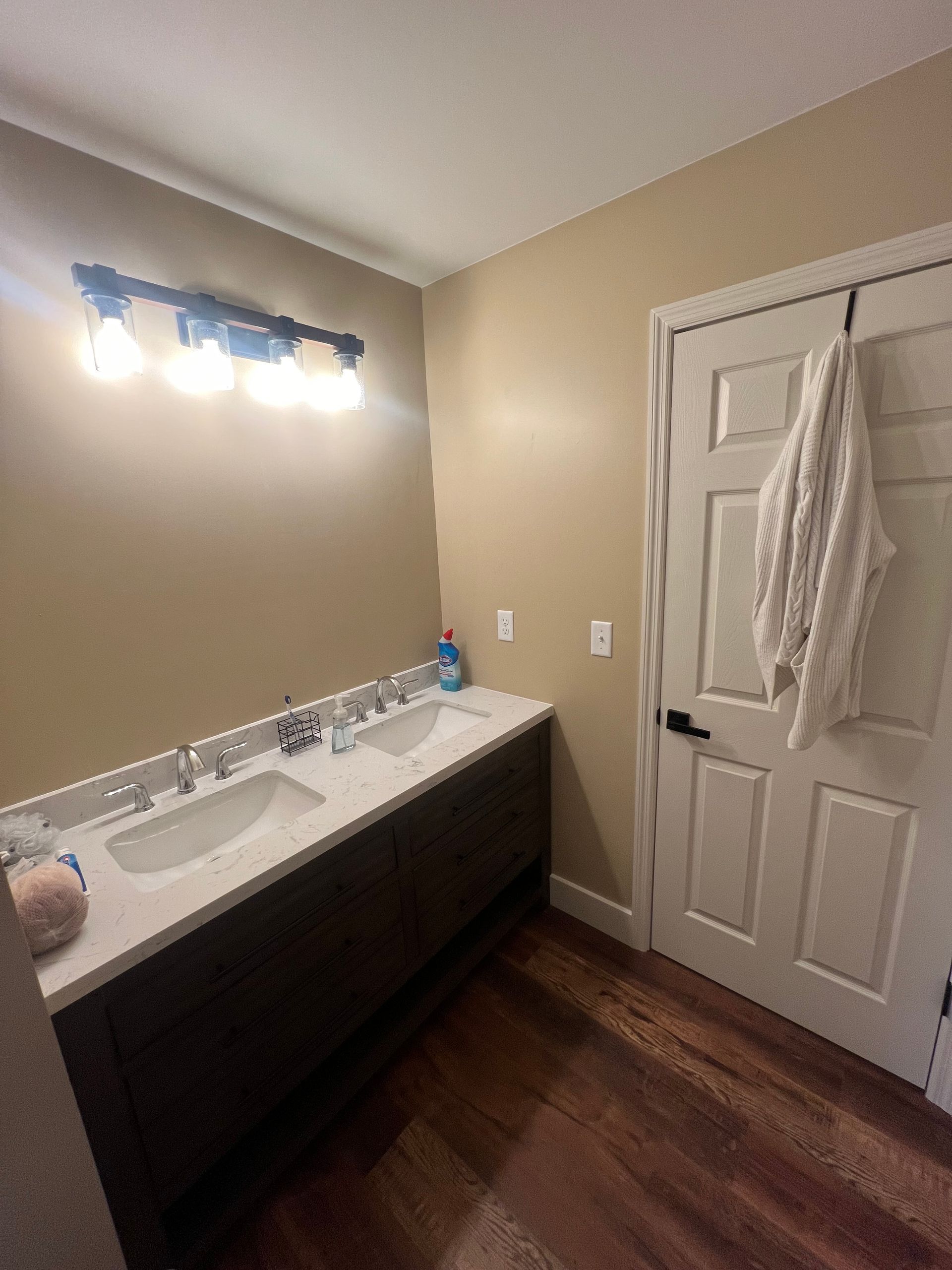 A bathroom with a double sink vanity, dark wood cabinets, and a light-colored countertop.  A white door with a towel hanging on it is to the right.