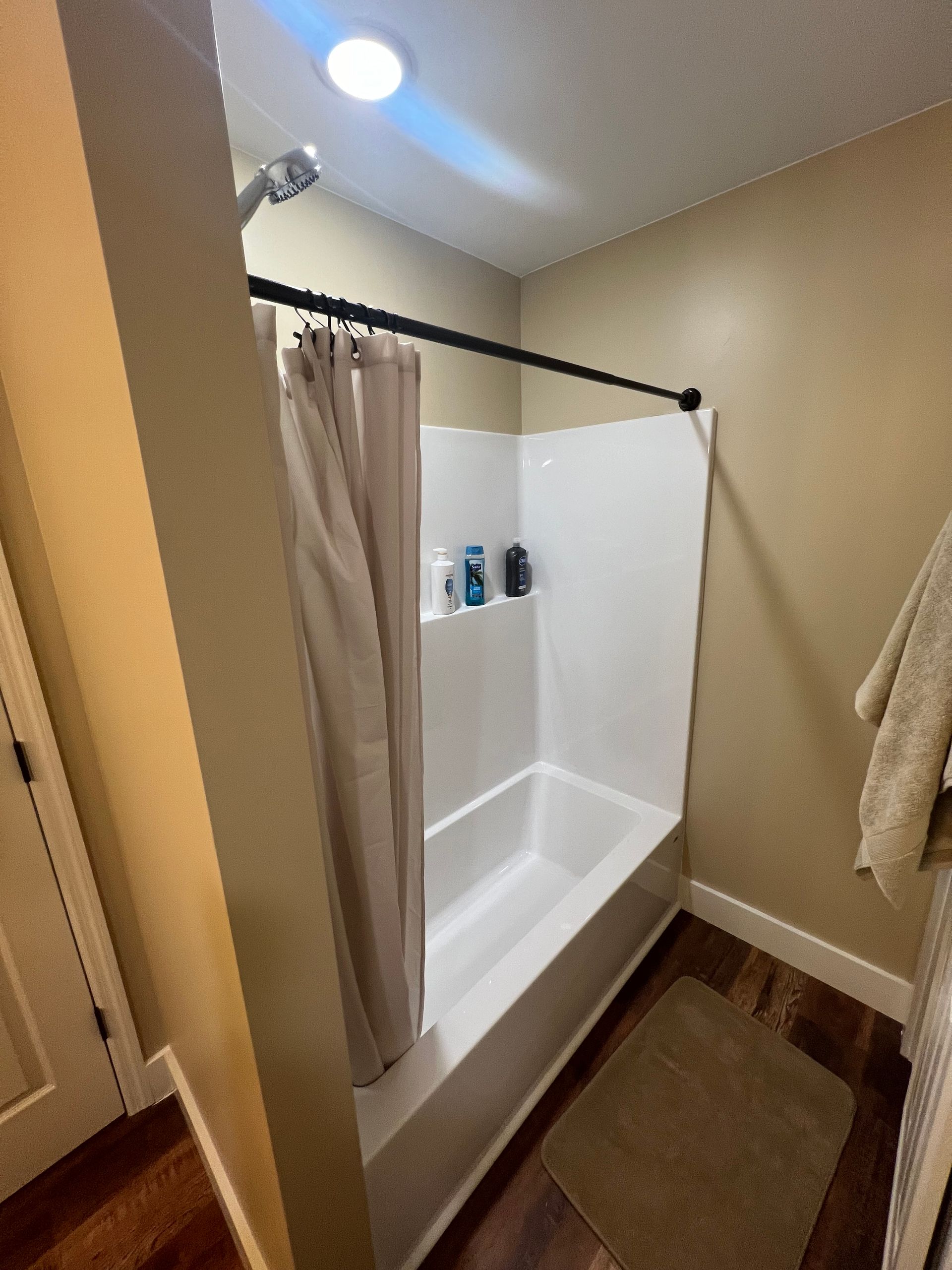 A small bathroom with a white shower/tub unit, beige walls, and a dark brown rug. A shower curtain hangs closed.