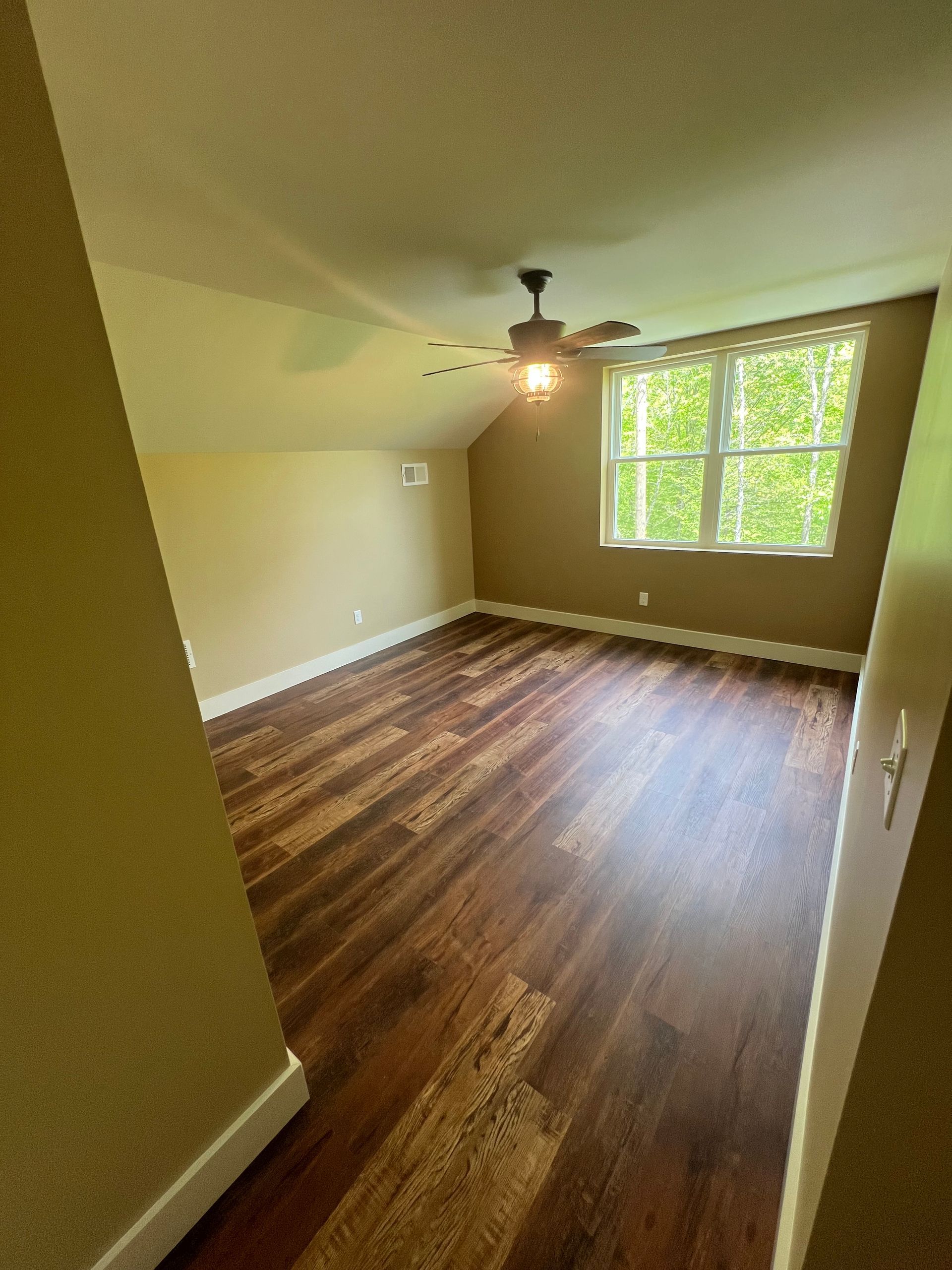 A room with dark wood flooring, a window with a view of green trees, and a ceiling fan. The walls are beige.