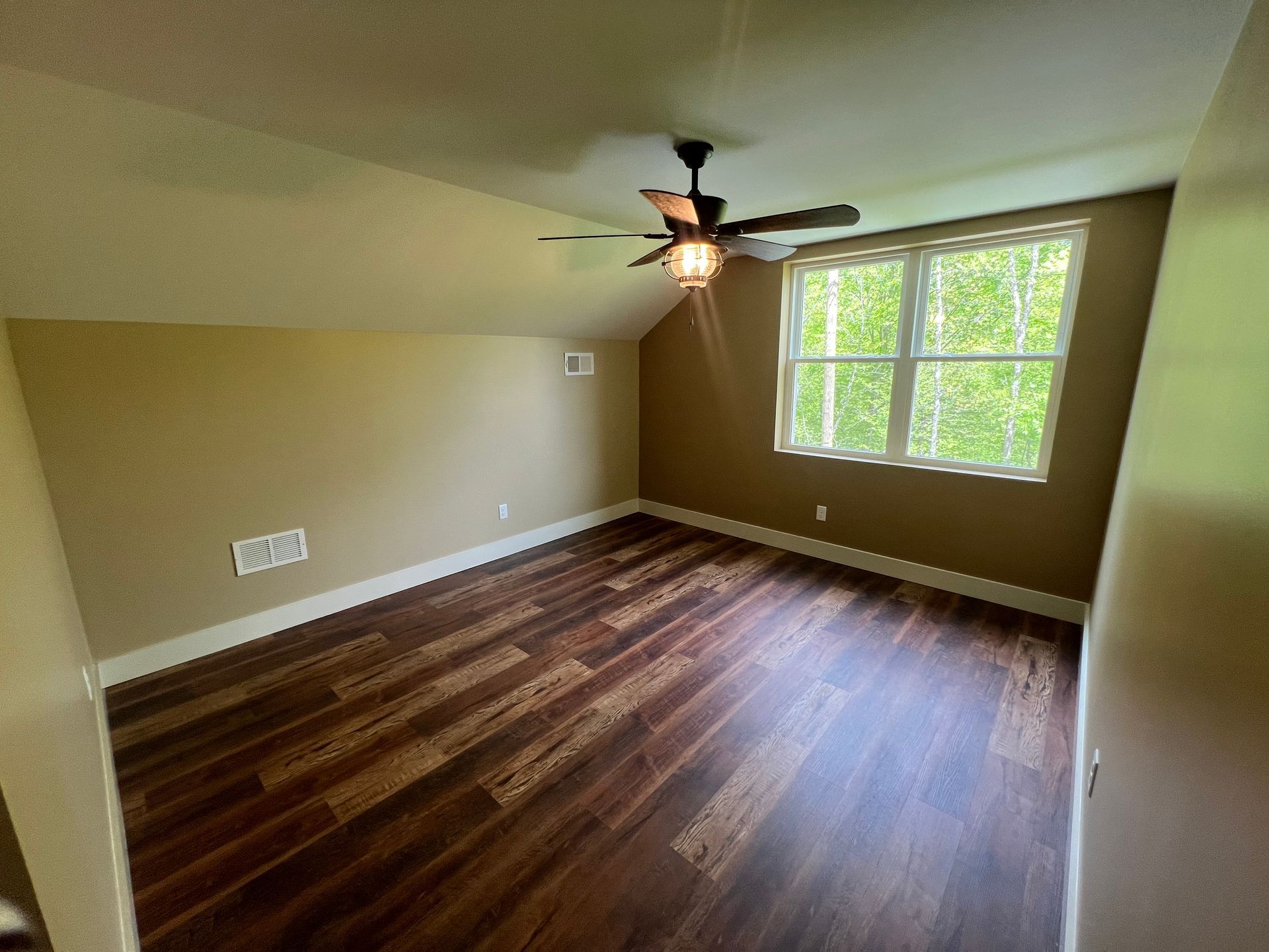 Bedroom with dark wood floor, beige walls, and a window overlooking trees. A ceiling fan hangs from the angled ceiling.