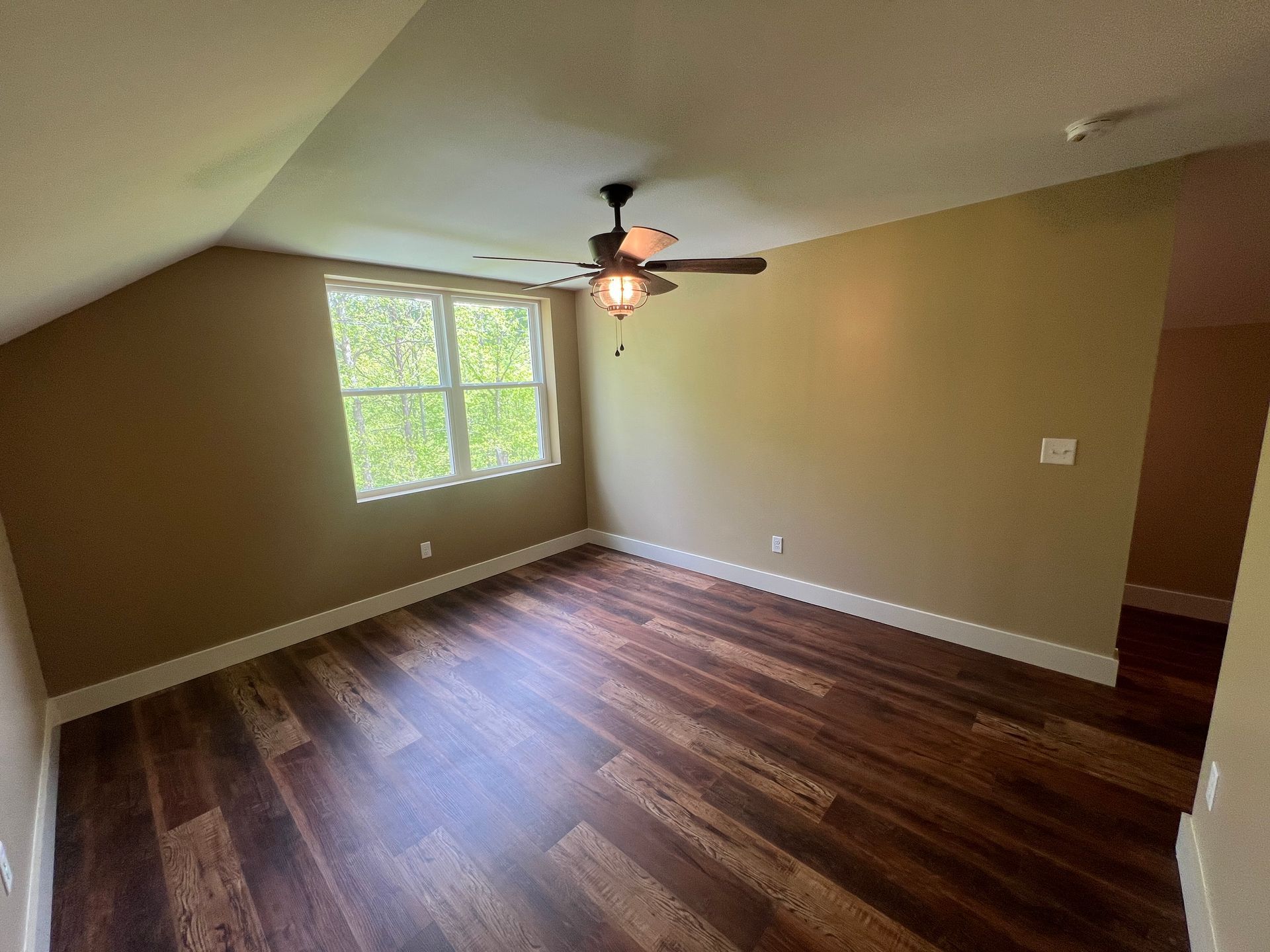 A sparsely furnished bedroom with dark wood floors, tan walls, a window, and a ceiling fan.