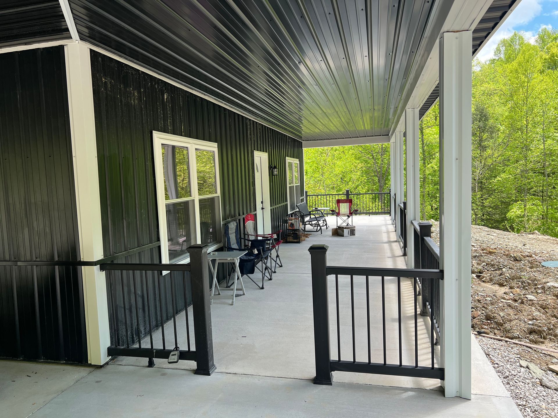 Black house with white trim, a covered porch with black railing, and surrounding green trees. Several folding chairs and tables are set up on the porch.