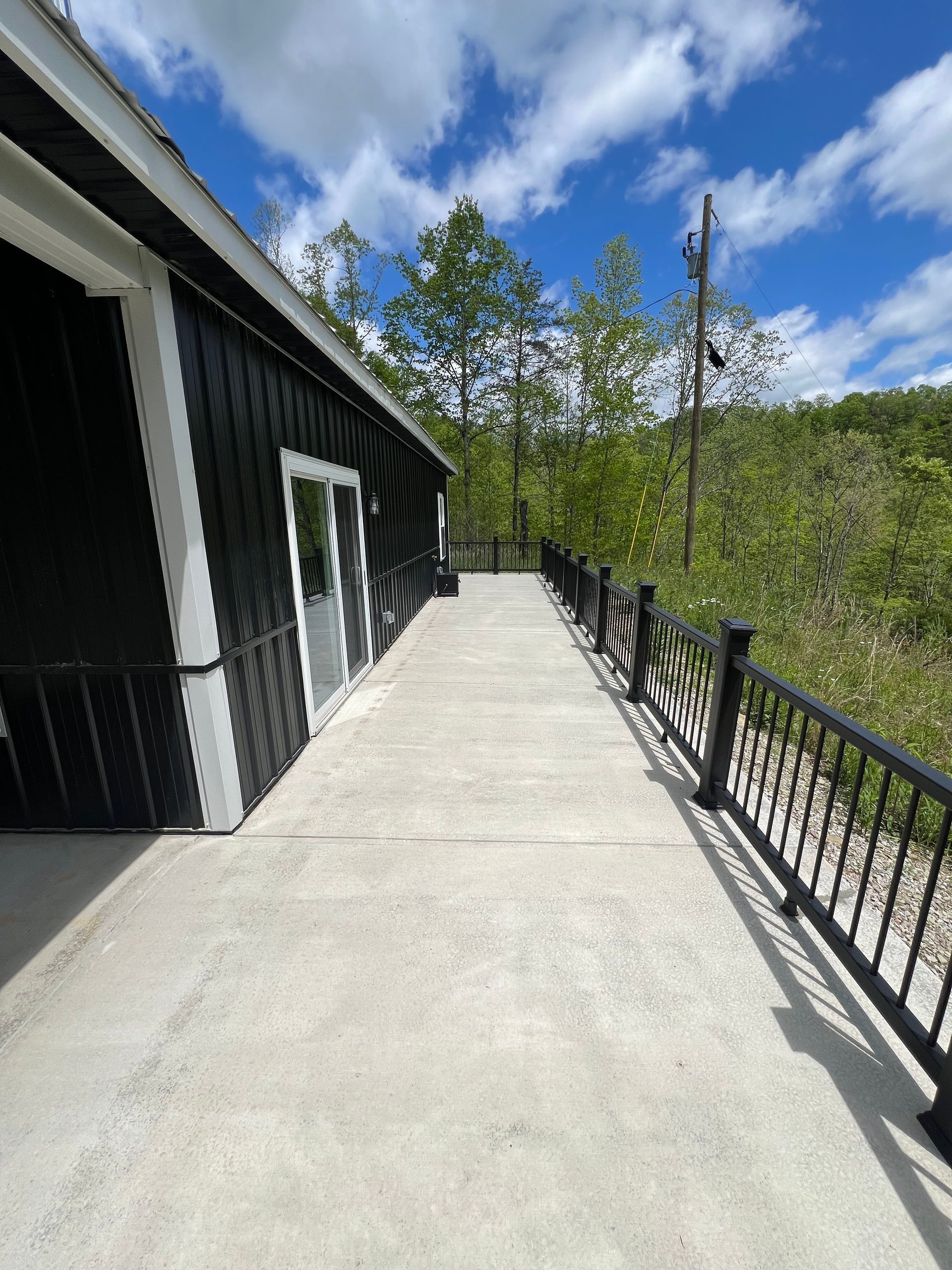 Black building with white trim and a concrete walkway with a black railing, set against a backdrop of trees and a partly cloudy blue sky.