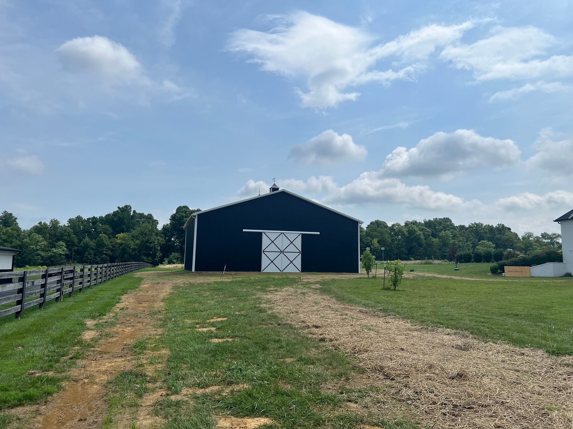 Black barn with white doors, set against a bright blue sky with scattered clouds, in a rural field setting.