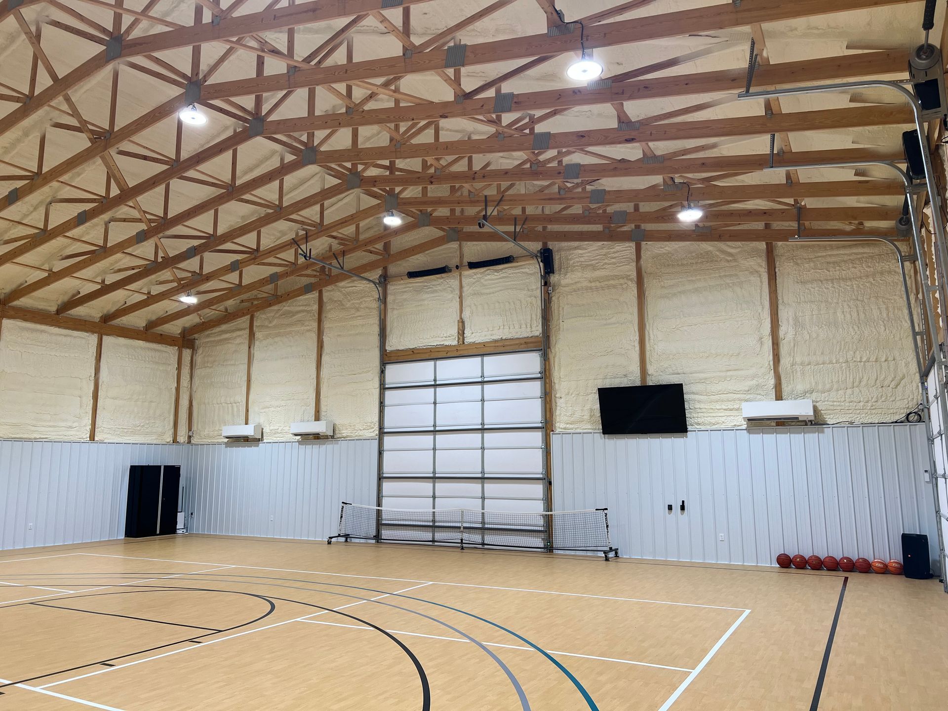 Basketball court interior with wooden beams, spray foam insulation, and a large garage door. The floor has lines for basketball.