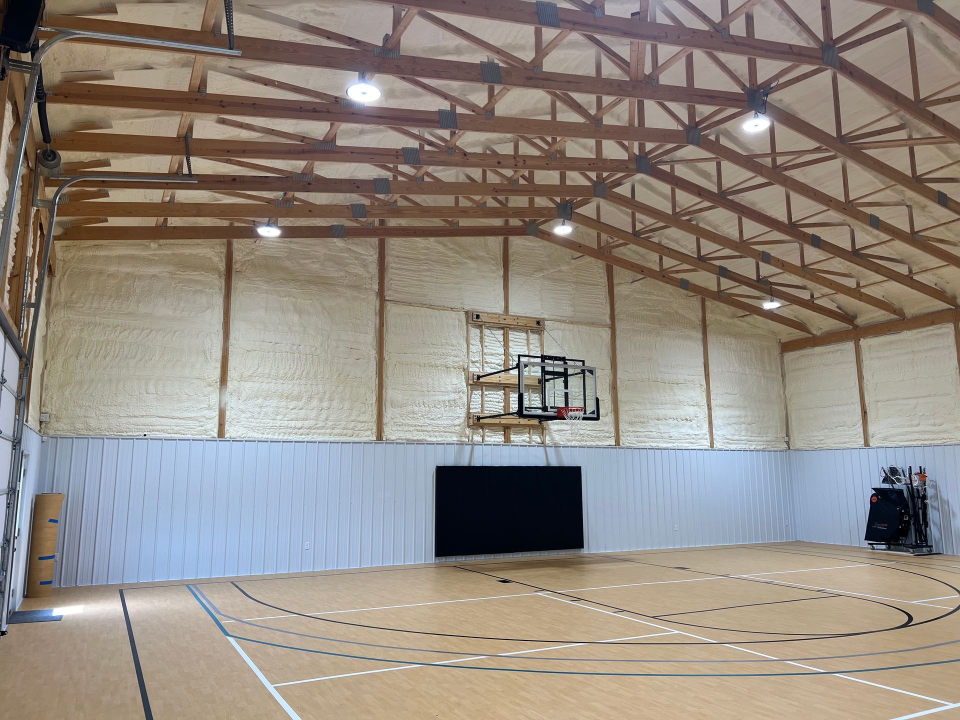 Interior view of a gymnasium with a basketball court, hoop, and insulation on the walls. The roof has exposed wooden beams.
