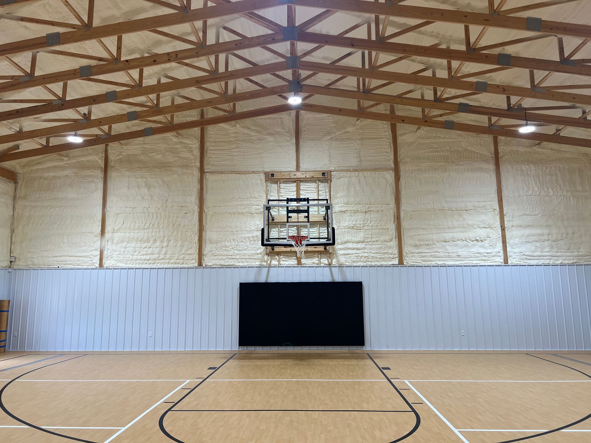 Interior of a basketball court. Wooden floor with black court lines, black scoreboard, and basketball hoop centered on the wall. Wooden beams on the ceiling.