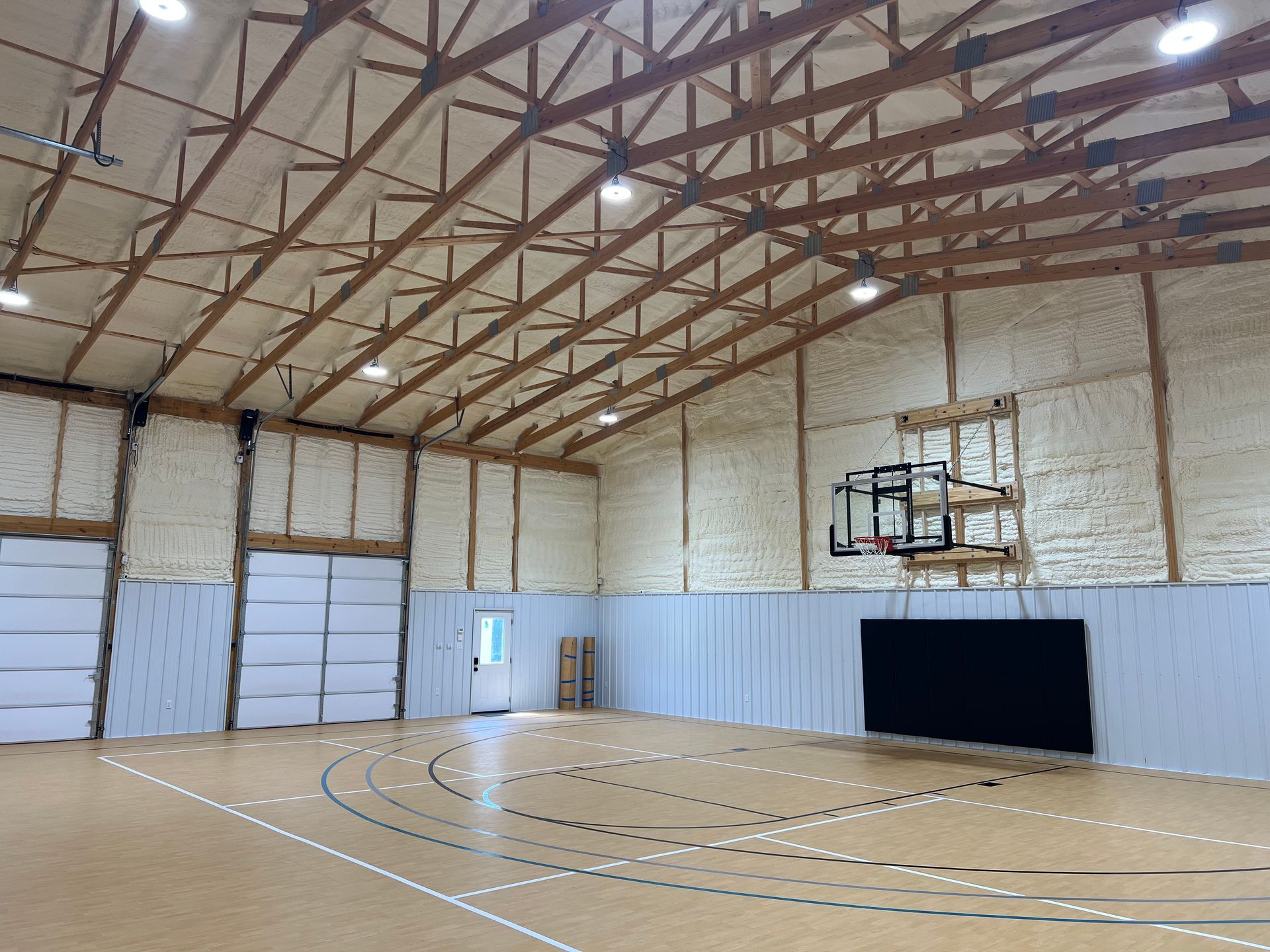Interior view of a gymnasium with a basketball hoop, wood truss ceiling, and basketball court markings. The walls and ceiling are covered in foam insulation.