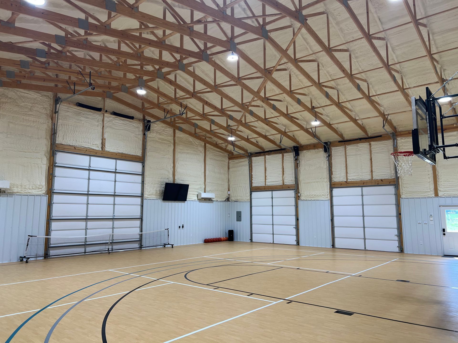 Interior of a gymnasium with a basketball hoop. There are three large white garage doors, brown beams, and a wood floor.