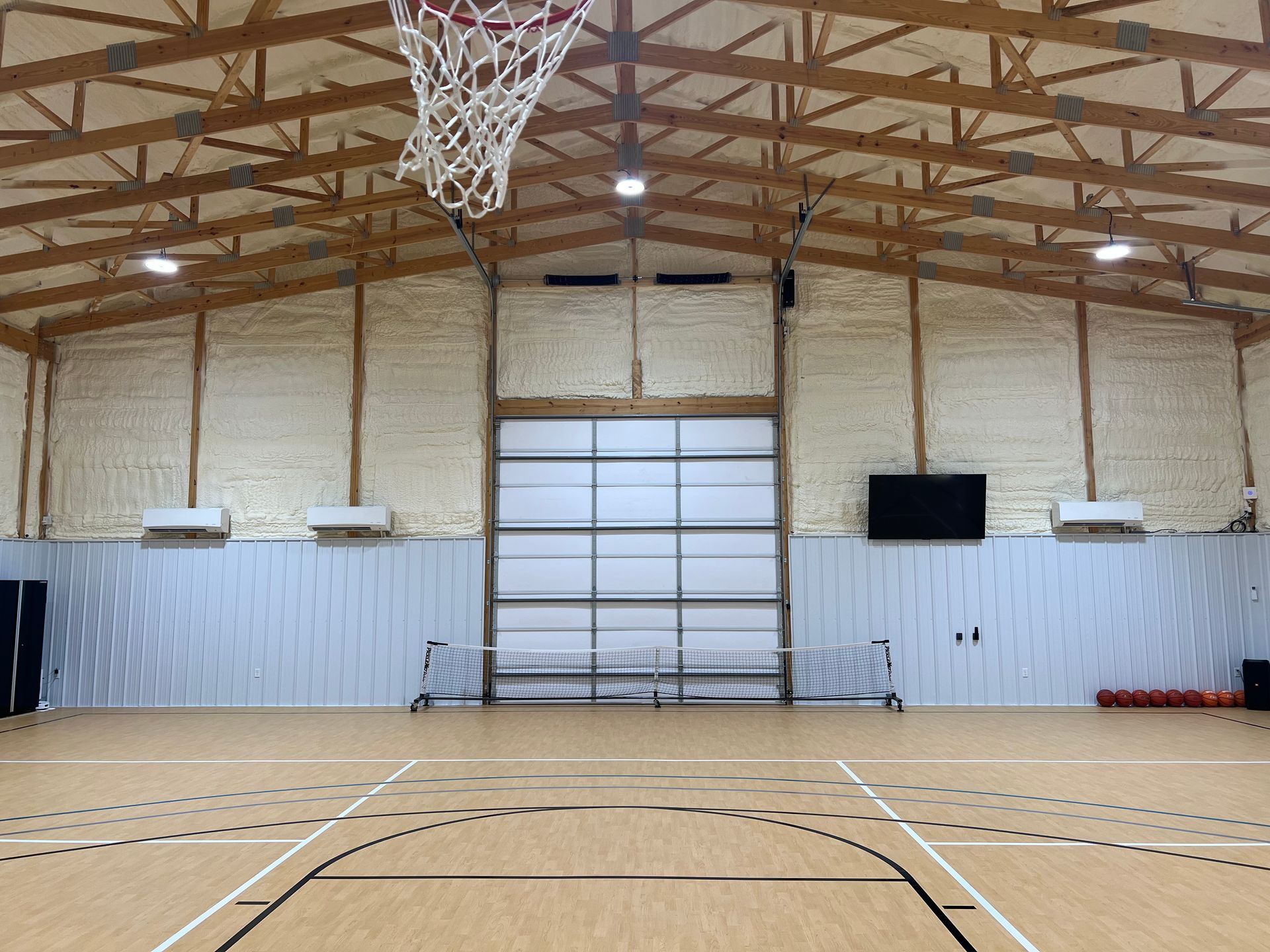 A gymnasium with a basketball court, hoop, and a closed garage door. The ceiling has exposed wooden beams.