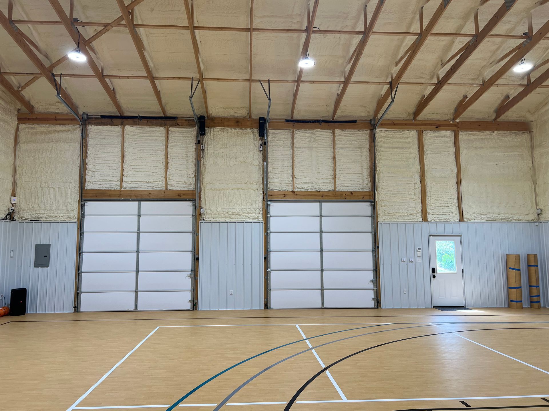 Interior of a gymnasium with hardwood floor and basketball court markings. Two large garage doors and a white door are visible. Insulation and exposed beams are on the walls and ceiling.