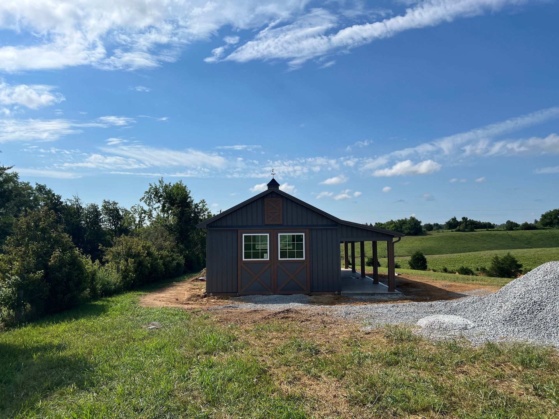A dark gray barn-like structure with a small porch in a grassy field under a blue sky.