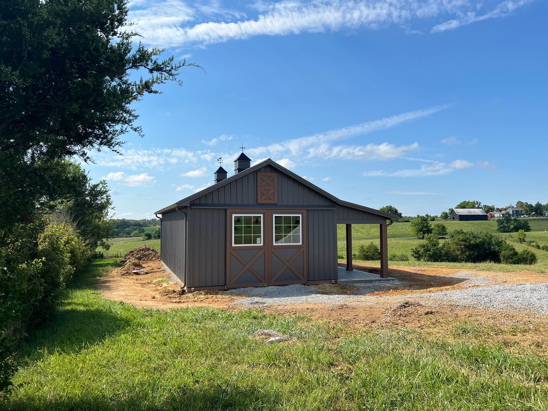A small, gray barn with two white-framed windows stands under a clear blue sky in a field.