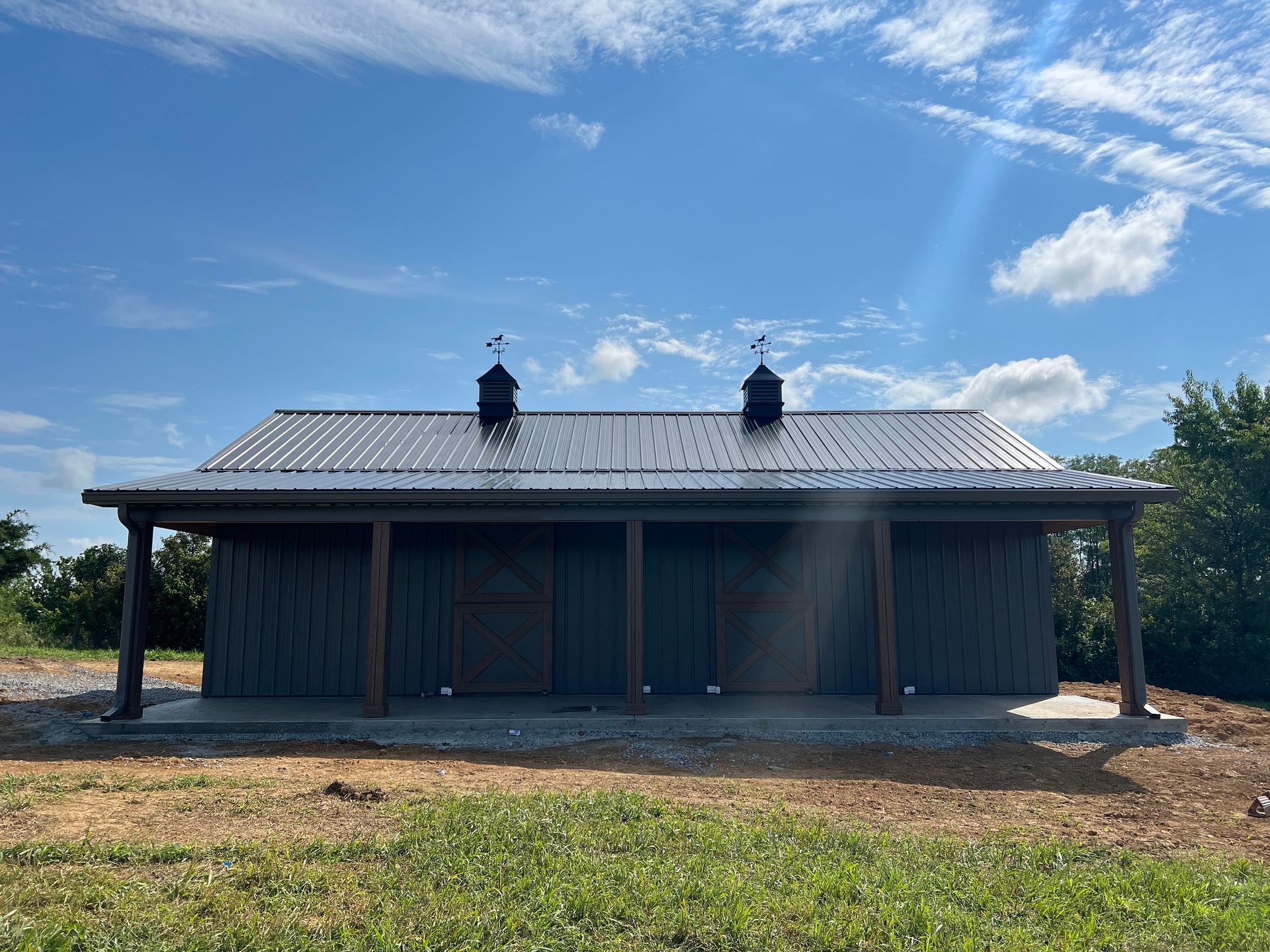 A dark gray barn with a metal roof under a blue sky with scattered clouds. Two stalls are visible under a covered porch.