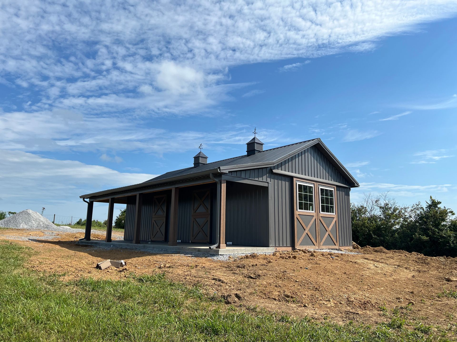 A dark gray barn with wooden accents and a covered porch stands under a blue sky.