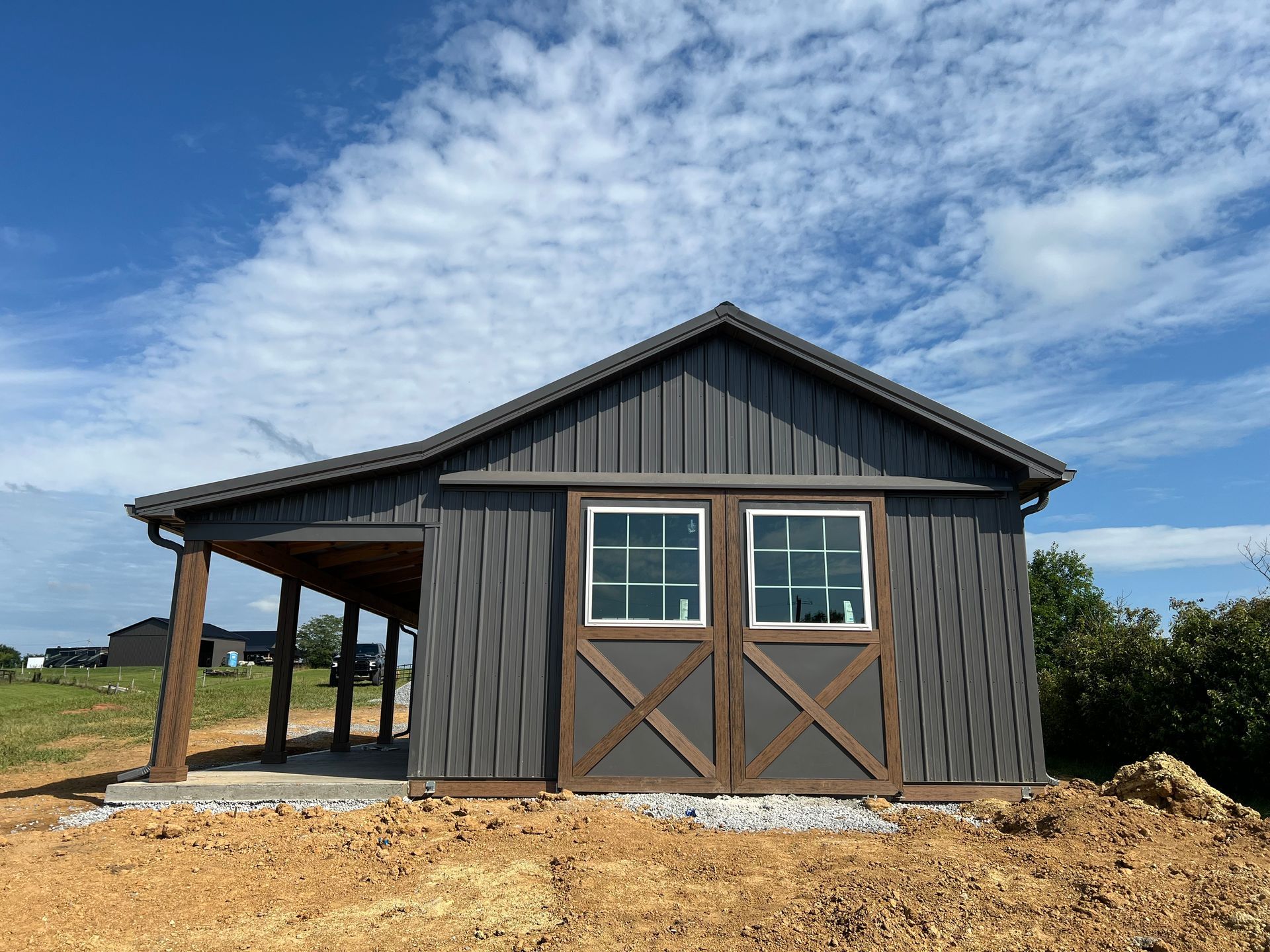 A gray barn with brown sliding doors, windows, and a carport, set against a blue sky and dirt ground.