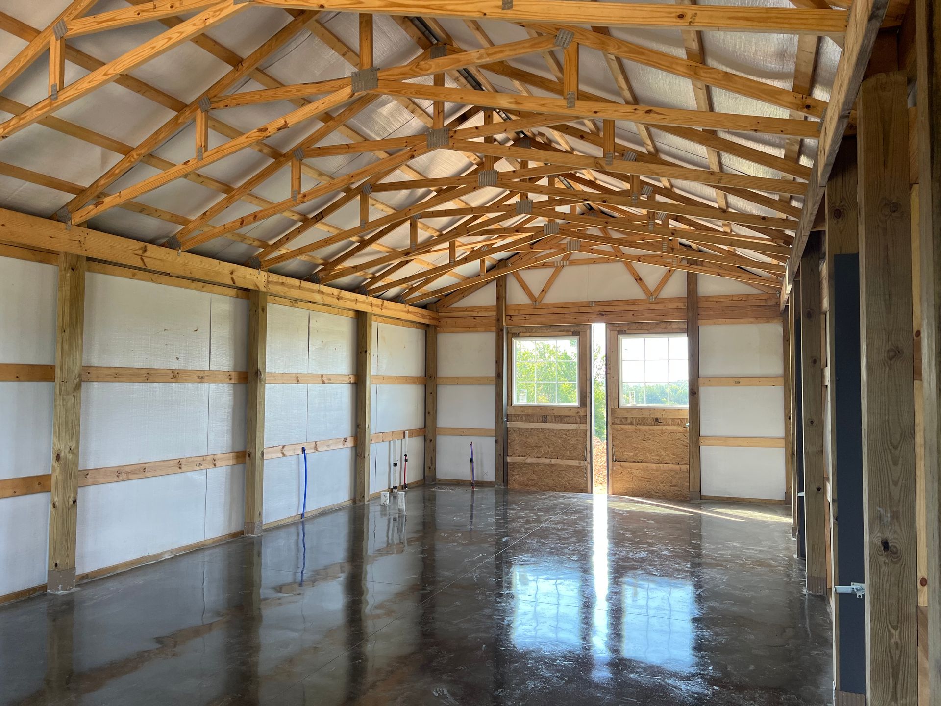 Interior view of an unfinished wooden structure with a concrete floor, showing exposed beams and rafters. Sunlight streams through double doors.
