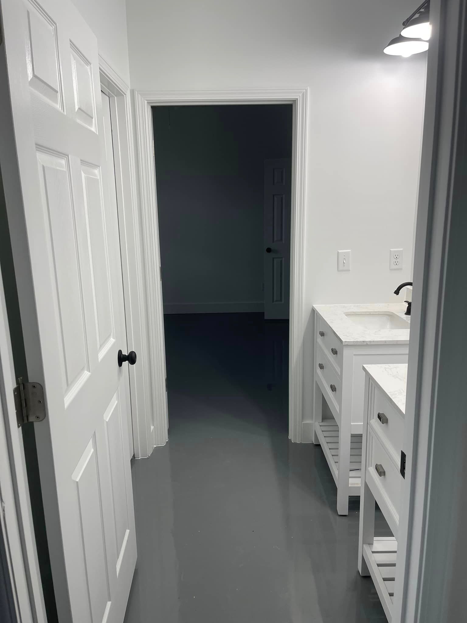 A narrow hallway with a white door on the left and two white vanities on the right. The floor is gray.