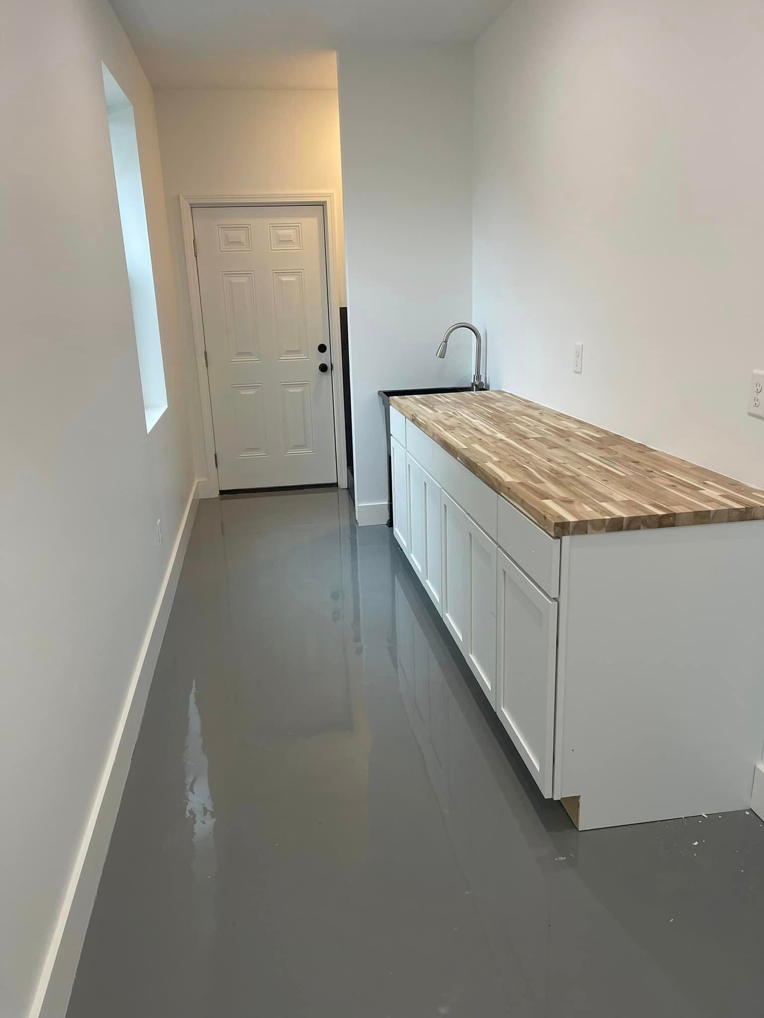 A narrow utility room with a long white cabinet and sink. A white door is at the end, and the floor is gray.