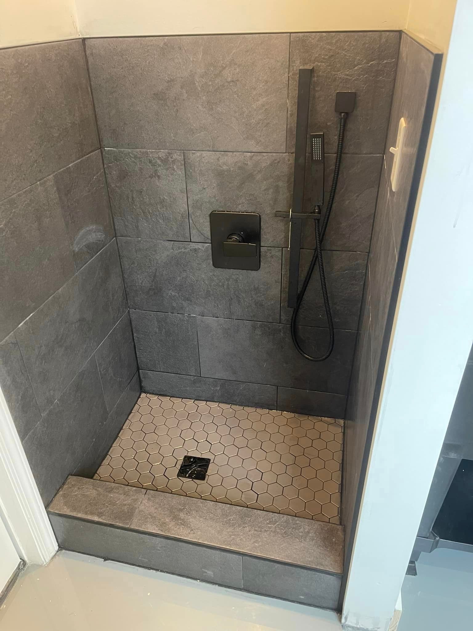 A small shower with gray stone tile walls and a pebbled floor. The showerhead and controls are black.