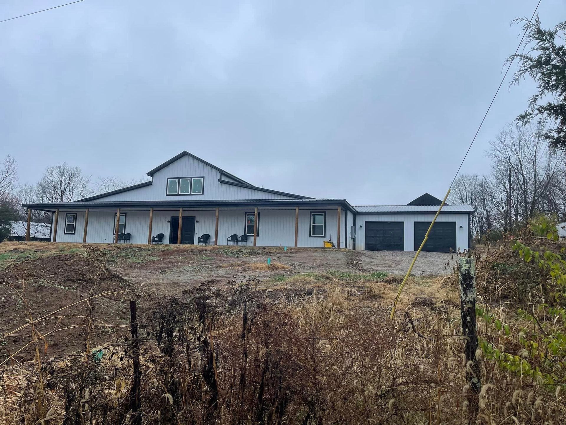 White house with black trim, connected to a garage, sits on a hillside under a cloudy sky.