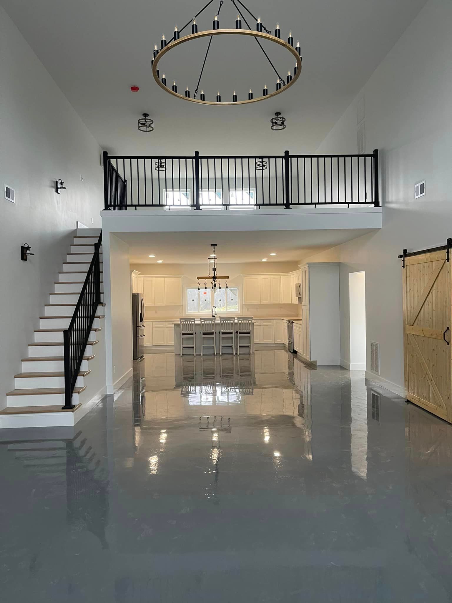 An open-concept home with a loft, gray floors, and a large chandelier above the kitchen island. Stairs lead to the loft, with a barn door on the right.