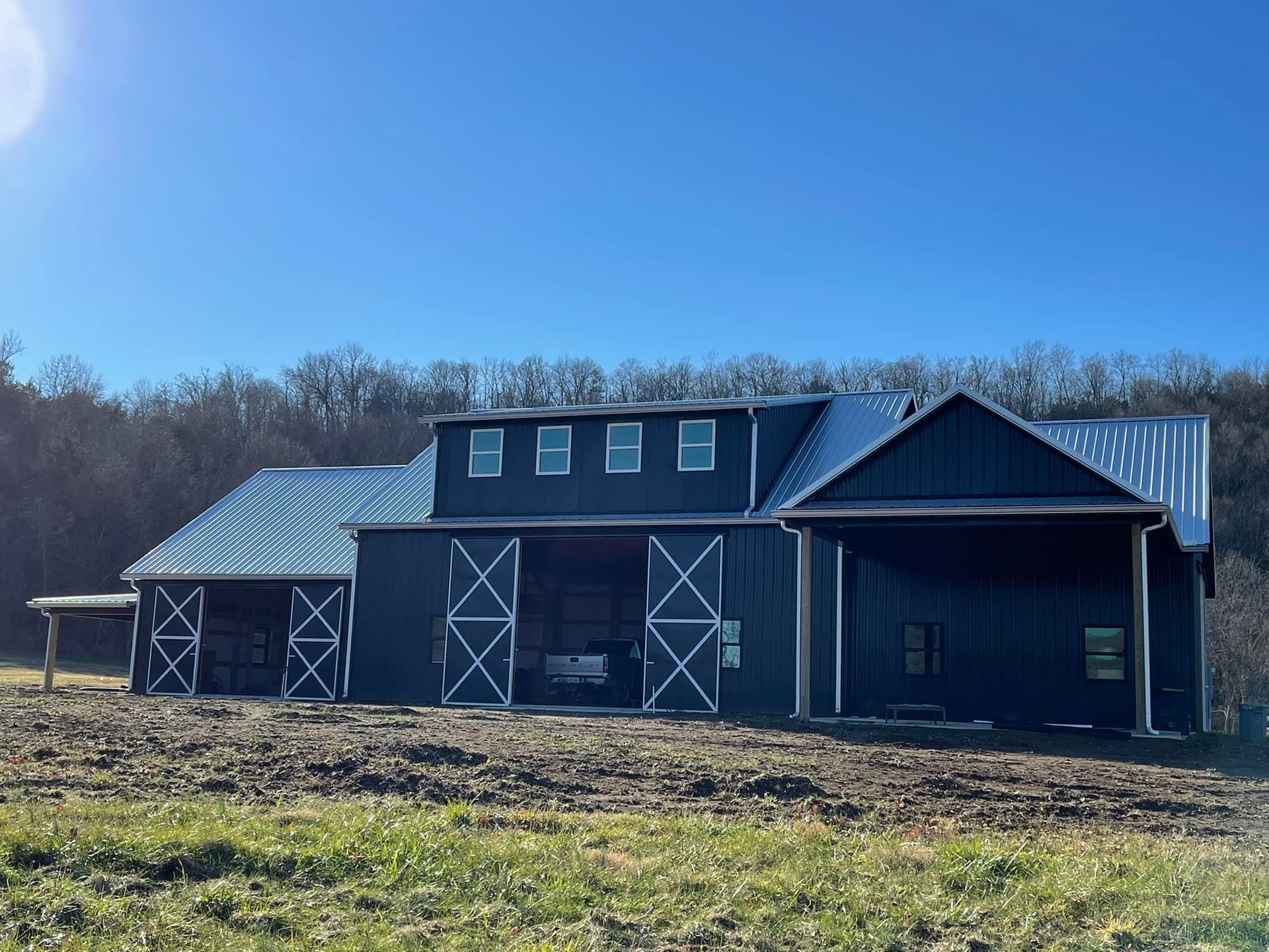 A large, dark blue barn with a metal roof sits in a field under a clear, blue sky.  The barn has open doors and windows.