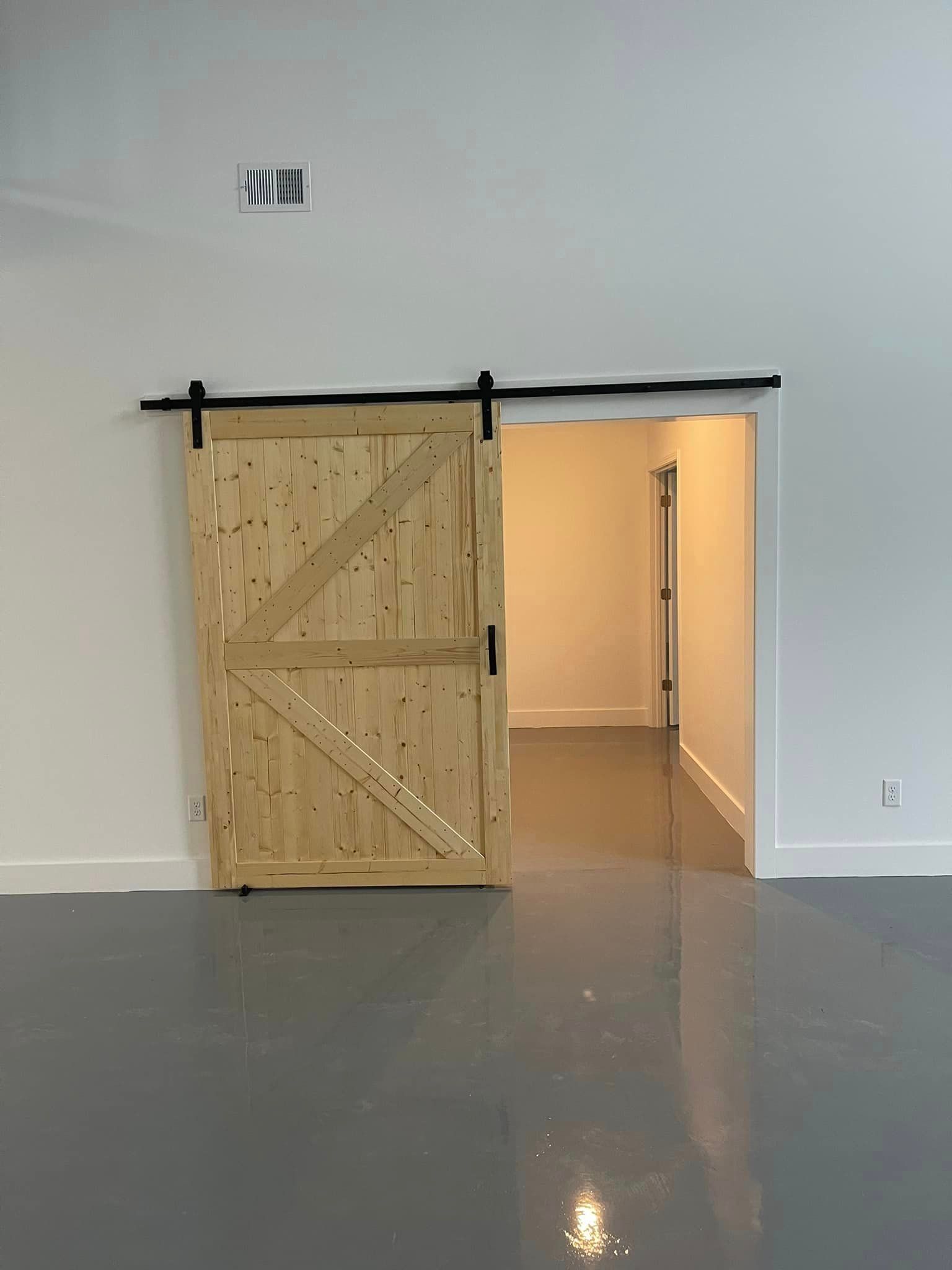 Light-colored wooden sliding barn door, with black hardware, open to a neutral room with a gray floor.
