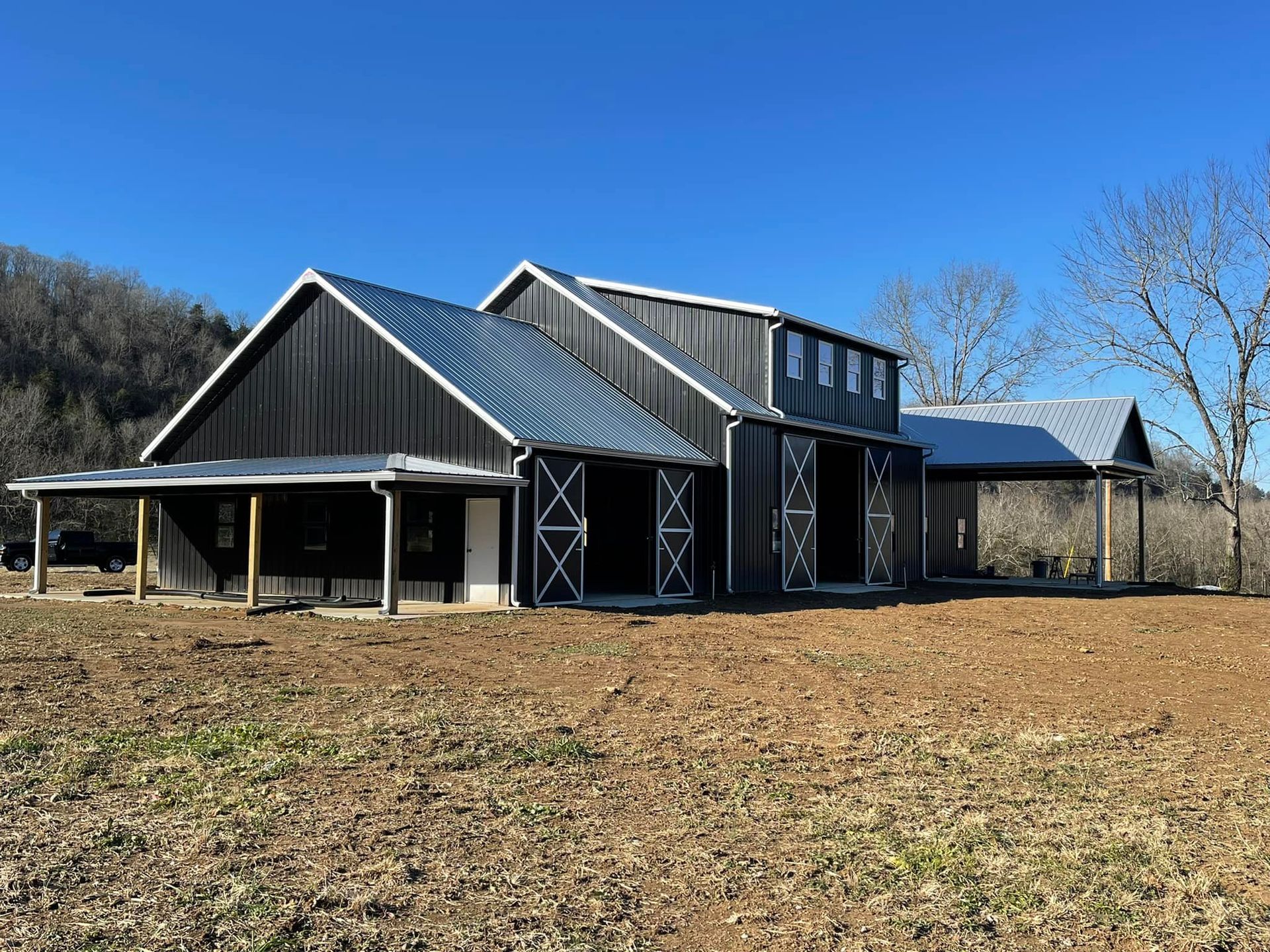 Dark gray barn with a metal roof against a clear blue sky. Open doors reveal the interior, and a porch extends from the side.