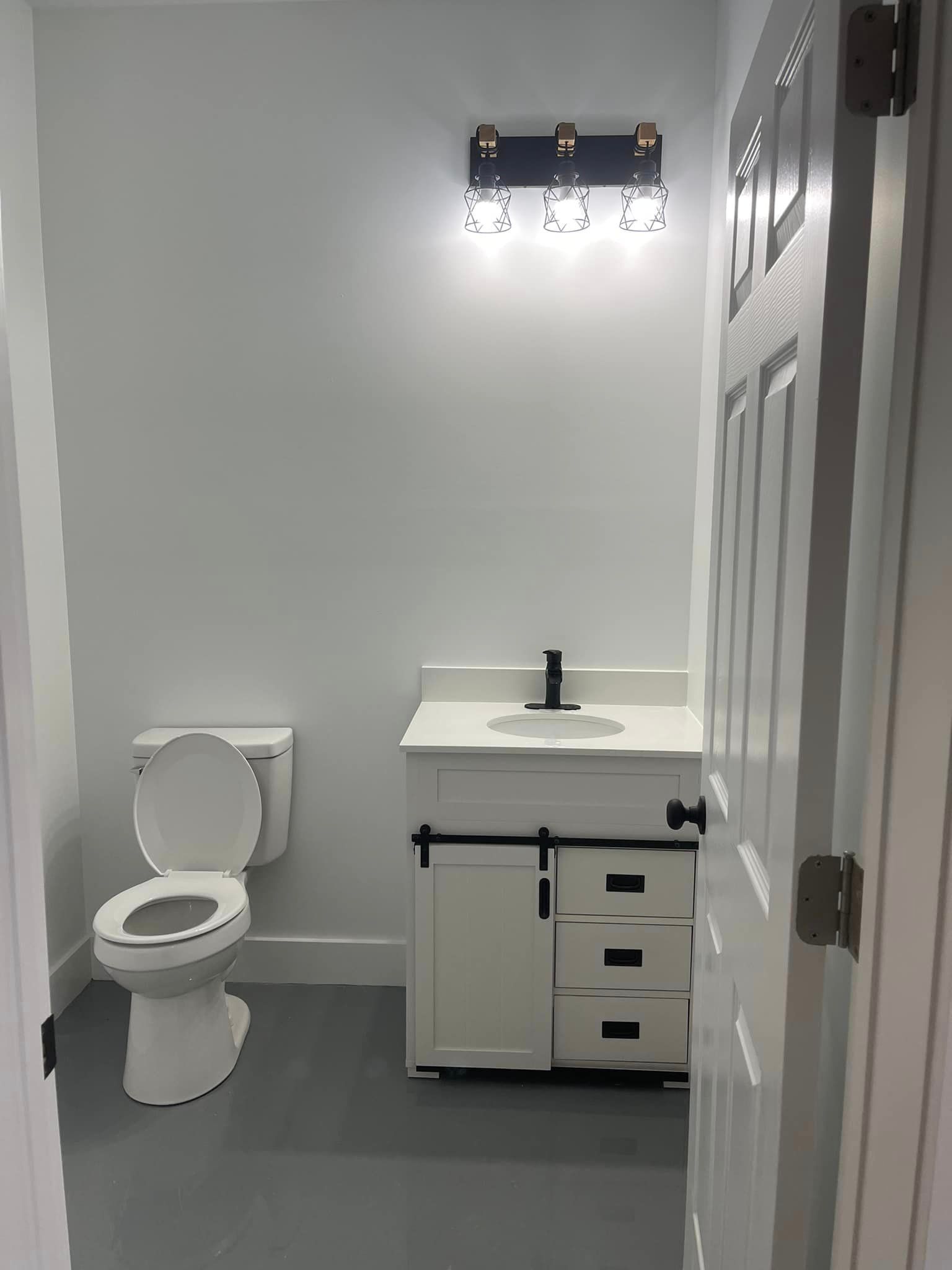 A small, white bathroom with a toilet, vanity, and door. Light fixture above the sink. The floor is gray.