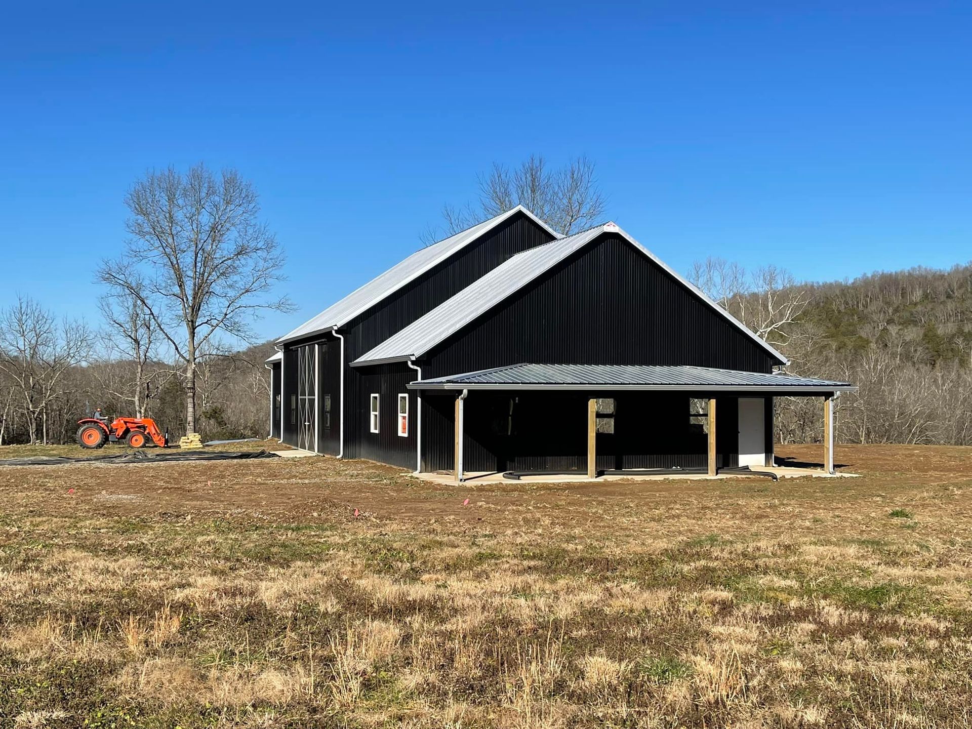 Black barn with a silver roof and front porch on a grassy field under a bright blue sky. A small orange tractor is parked in the yard.