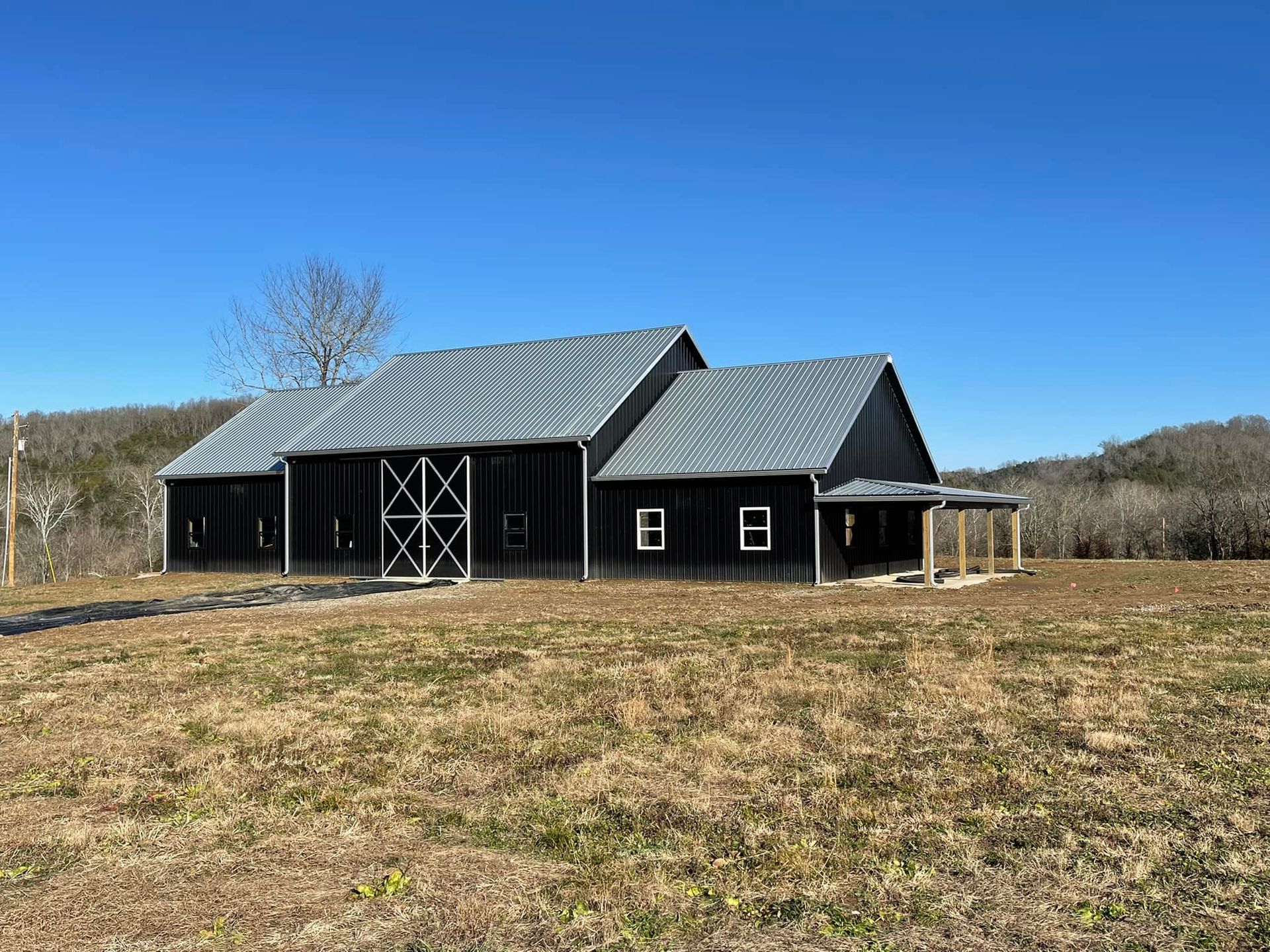 Black barn with a metal roof and white trim, sitting on a grassy hill under a clear blue sky.