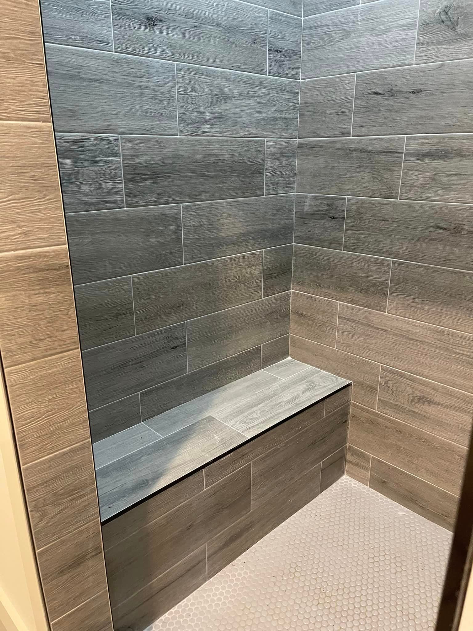 Shower stall with grey and brown tile and a built-in bench. The floor is covered in white hexagon tiles.