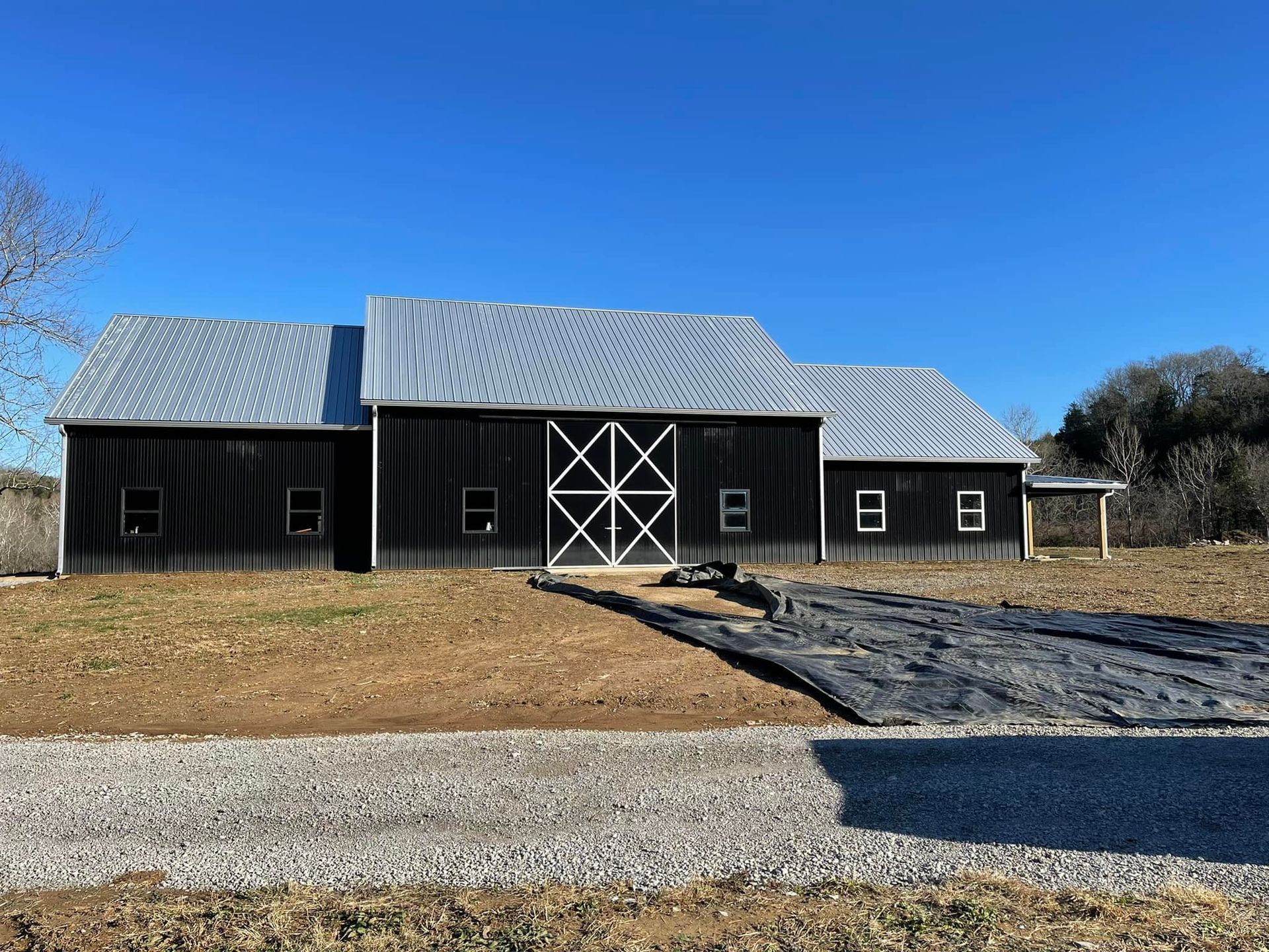 Black barn with a white-X door, silver roof, and gravel driveway under a blue sky.