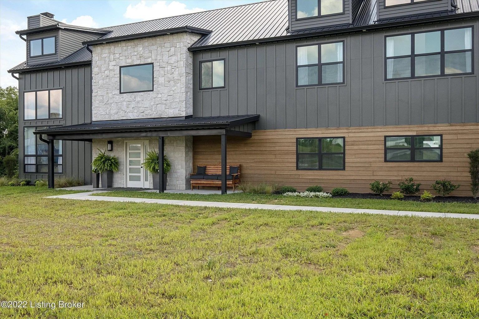 Modern two-story house with gray siding, stone accent wall, and wooden porch. Green lawn in front.
