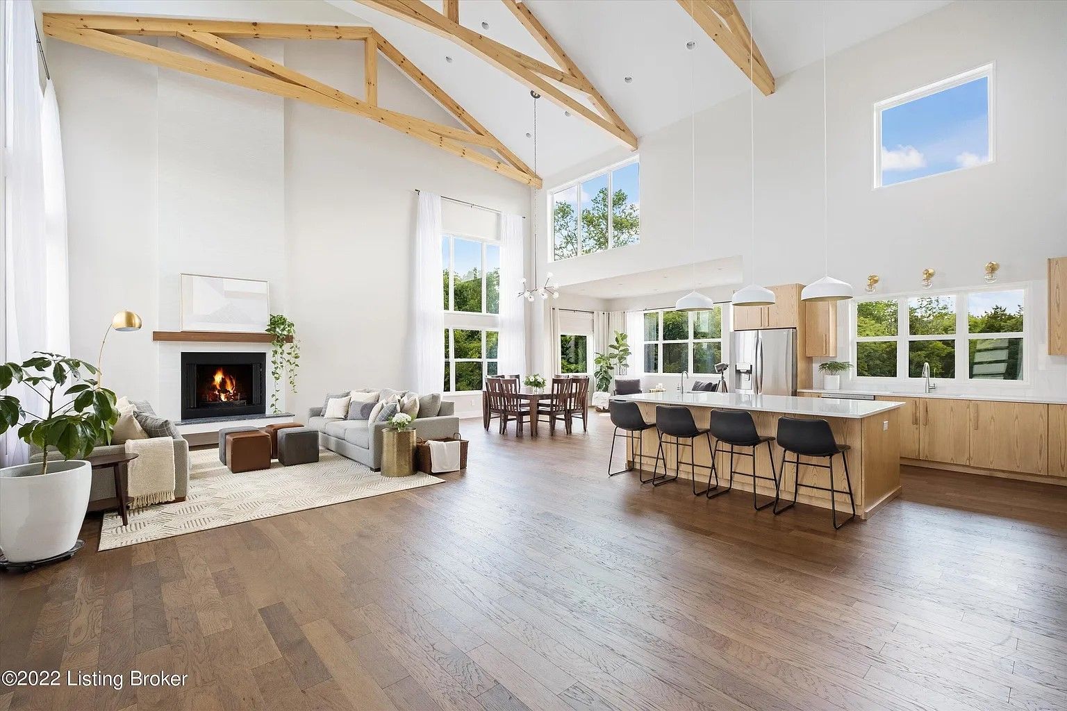 Spacious, light-filled living space with a high, exposed beam ceiling. Includes a fireplace, kitchen with island seating, and large windows.