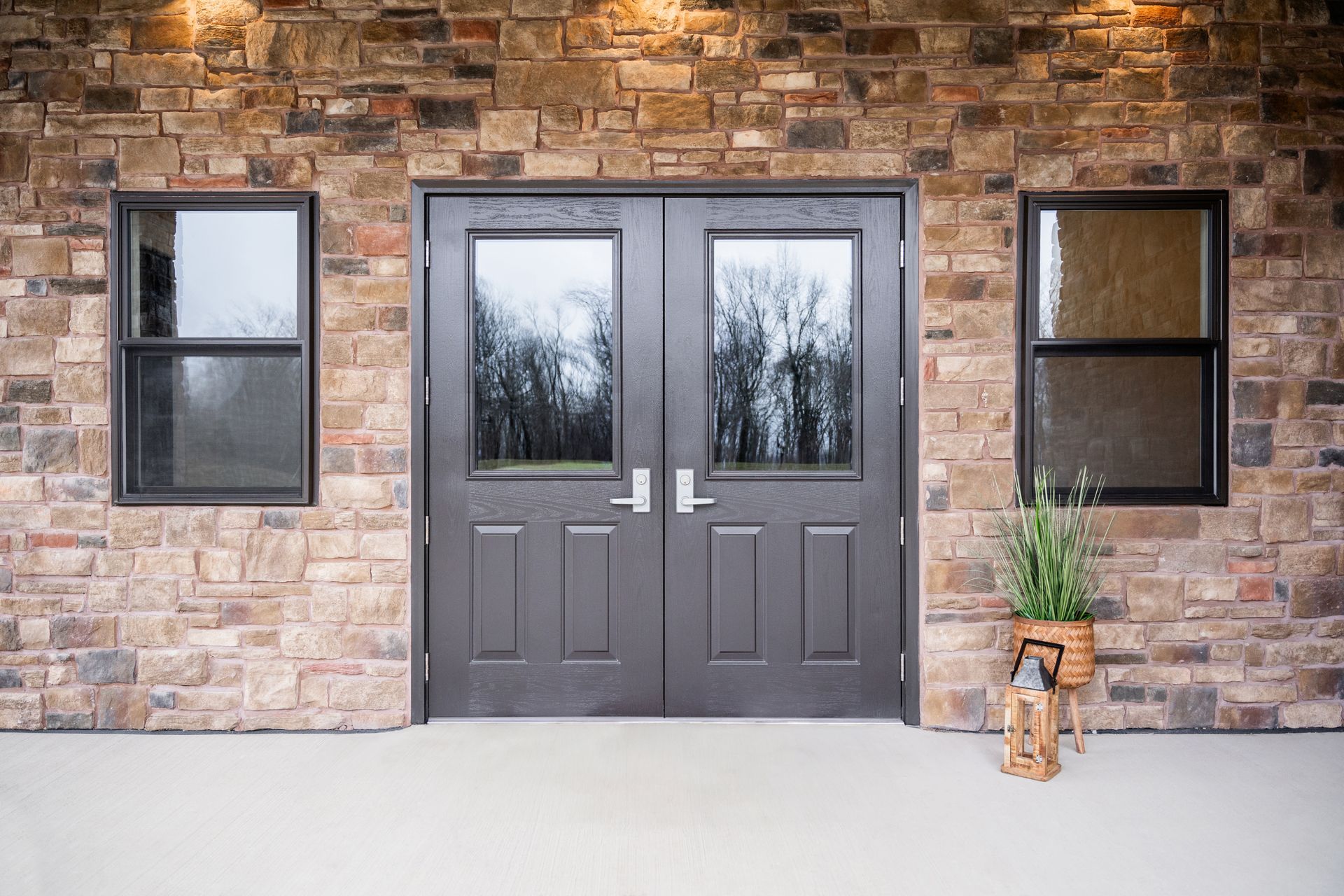 Exterior of a building with stone brick walls, a gray double door, two windows, and a small potted plant.