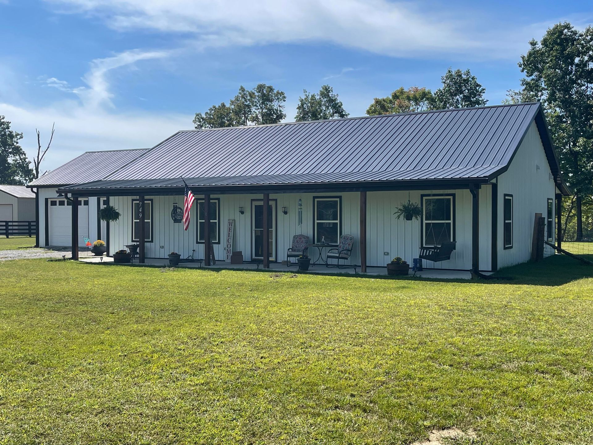 White farmhouse with a dark blue metal roof and porch, set on a grassy lawn under a blue sky.