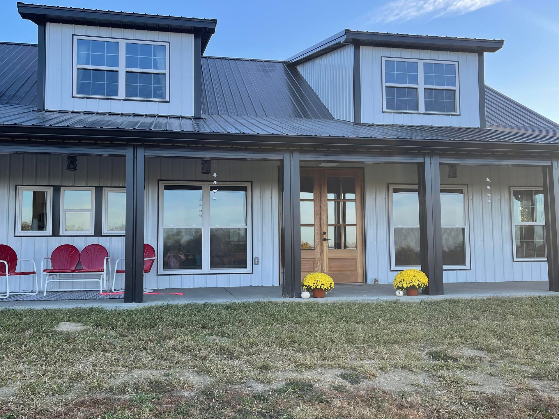 A farmhouse with a black metal roof and white siding. Two dormers and a front porch with a wooden door. Yellow sunflowers decorate the porch.