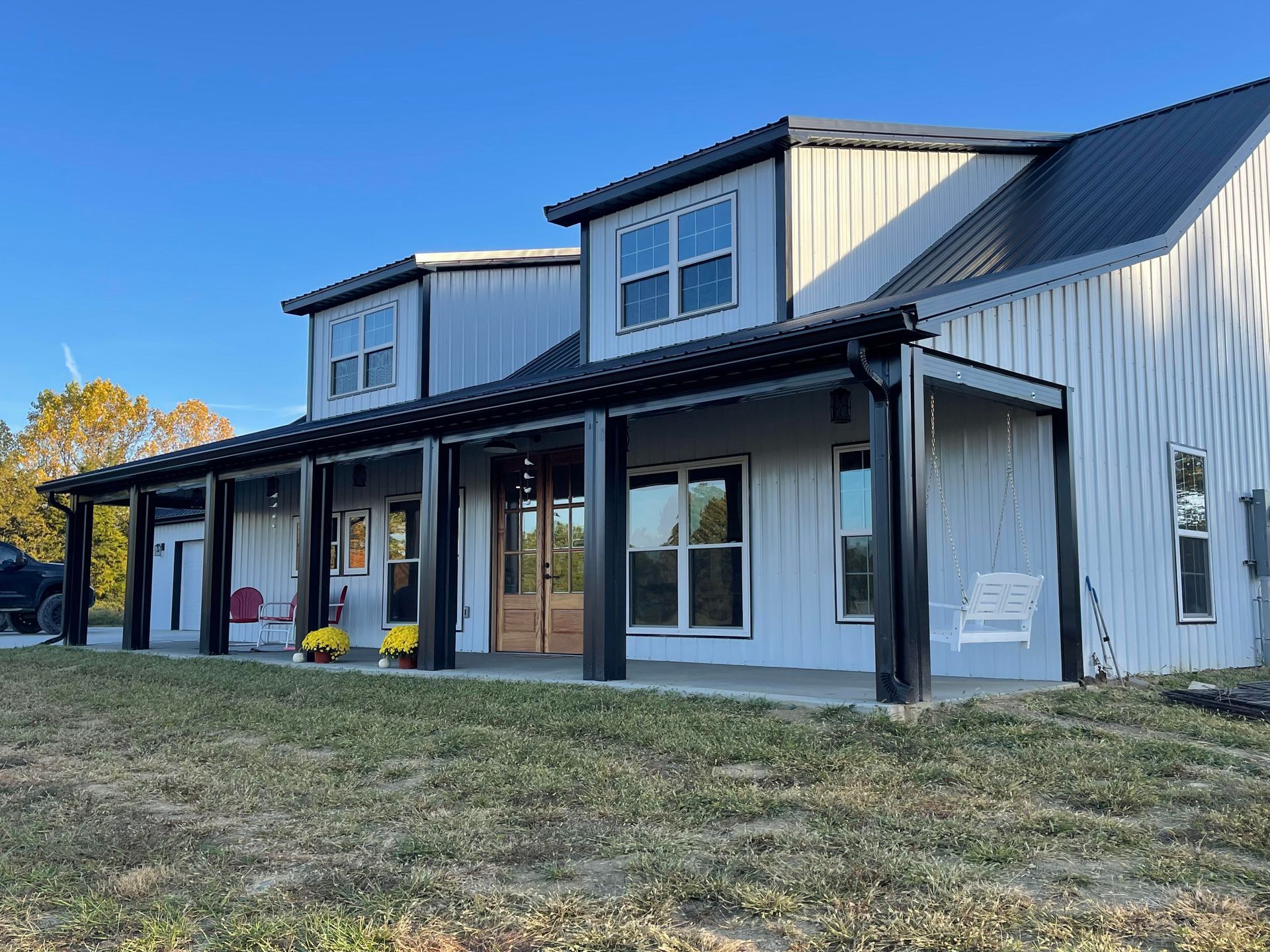 Two-story modern farmhouse with a black metal roof and porch.  The house is light gray with black trim and windows.  A grassy yard surrounds it.