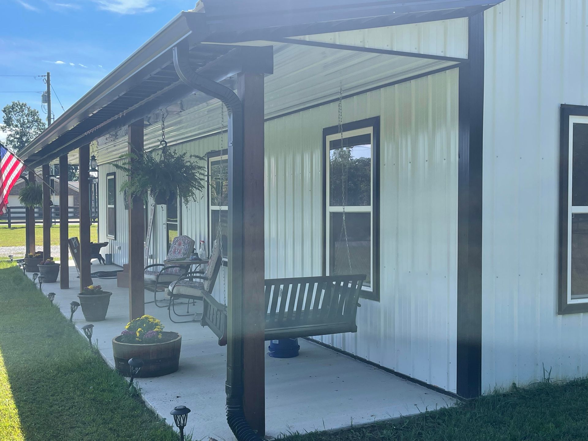 A white metal-sided house with a porch. A swing and seating are visible. The porch has brown posts and a dark metal roof.