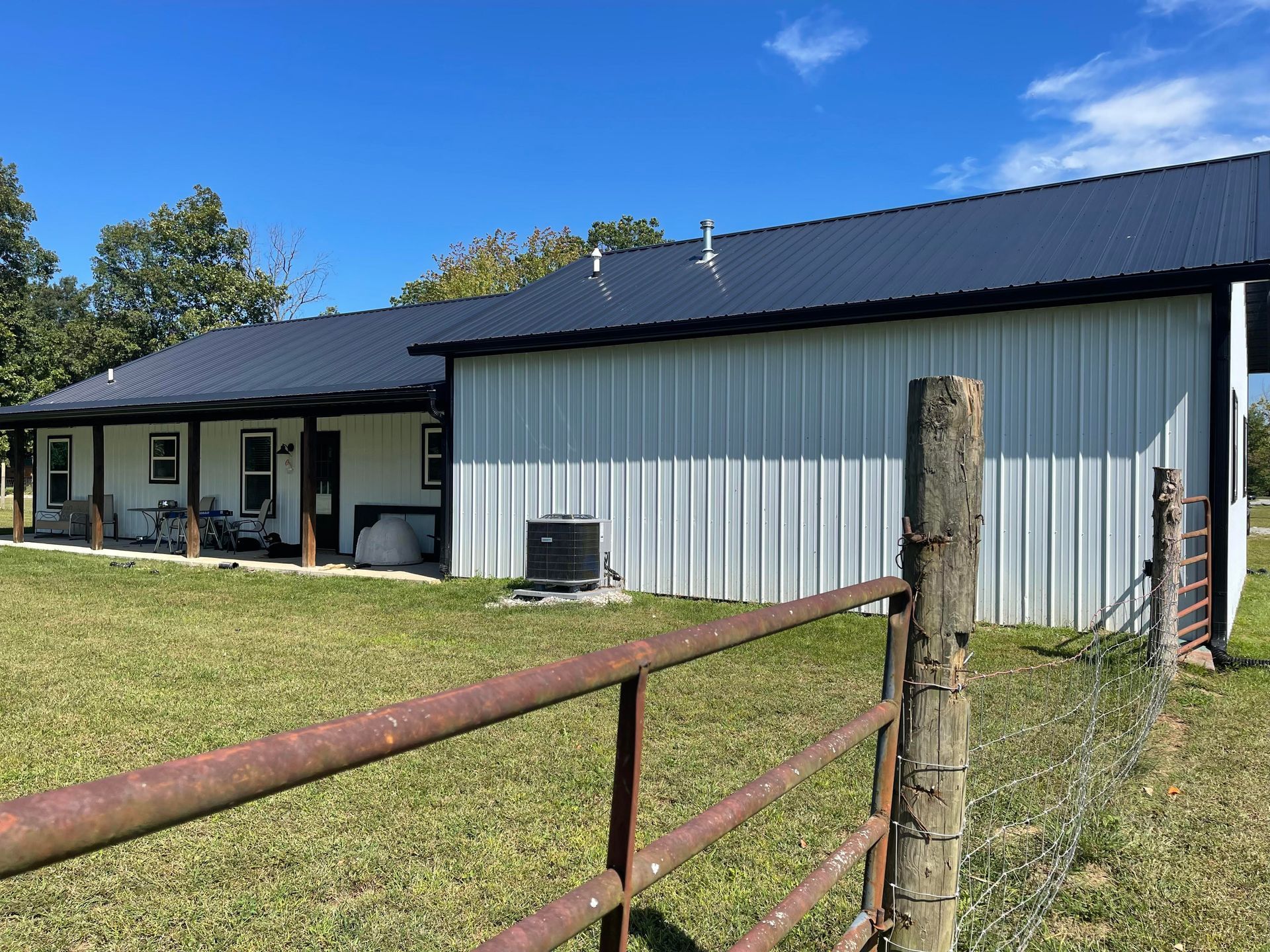 A long, white metal building with a dark blue roof and porch, viewed from a grassy field with a rusted fence.