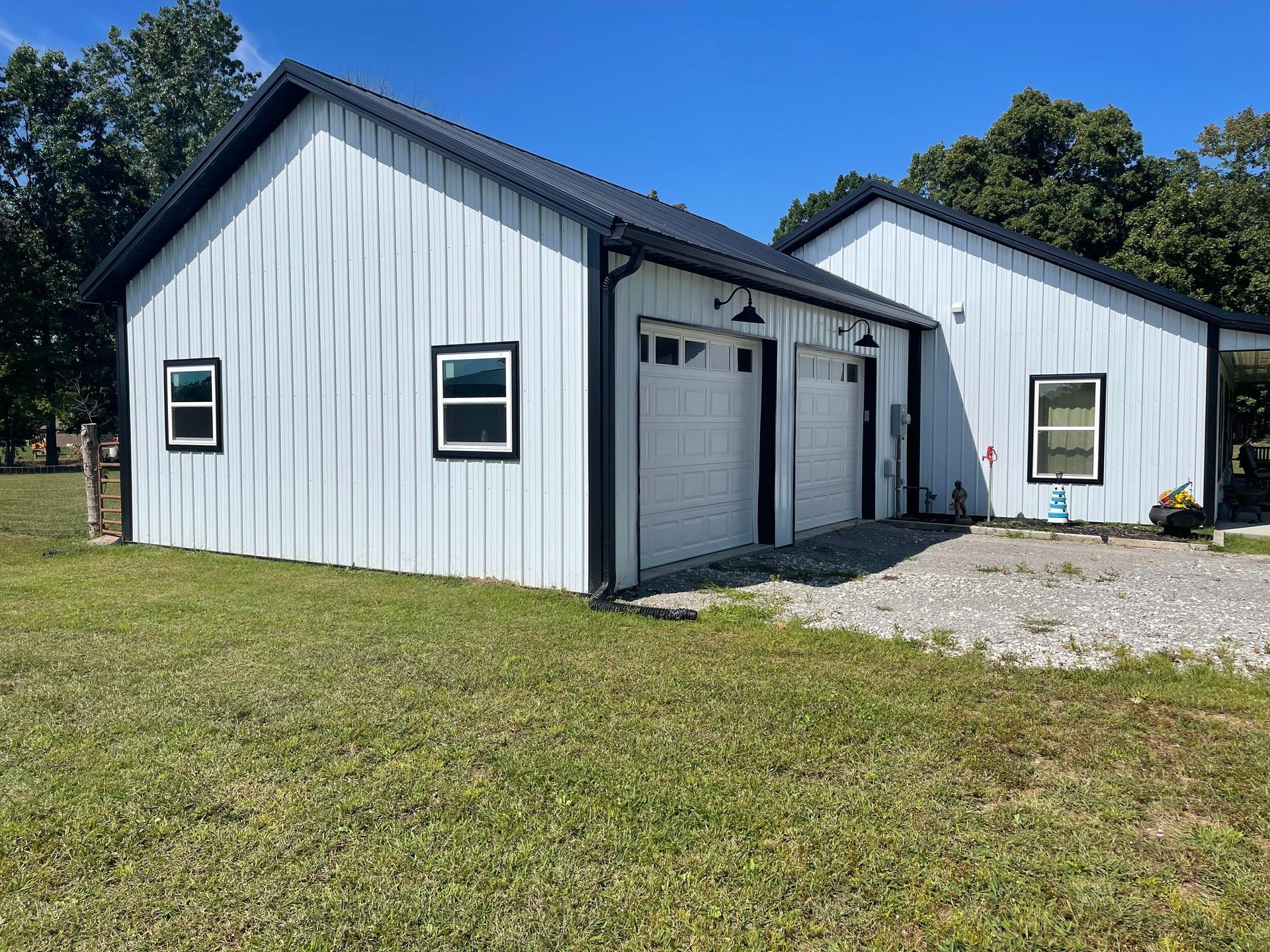 White barn-style building with black trim and roof. Features a two-car garage, windows, and is set in a grassy yard under a blue sky.