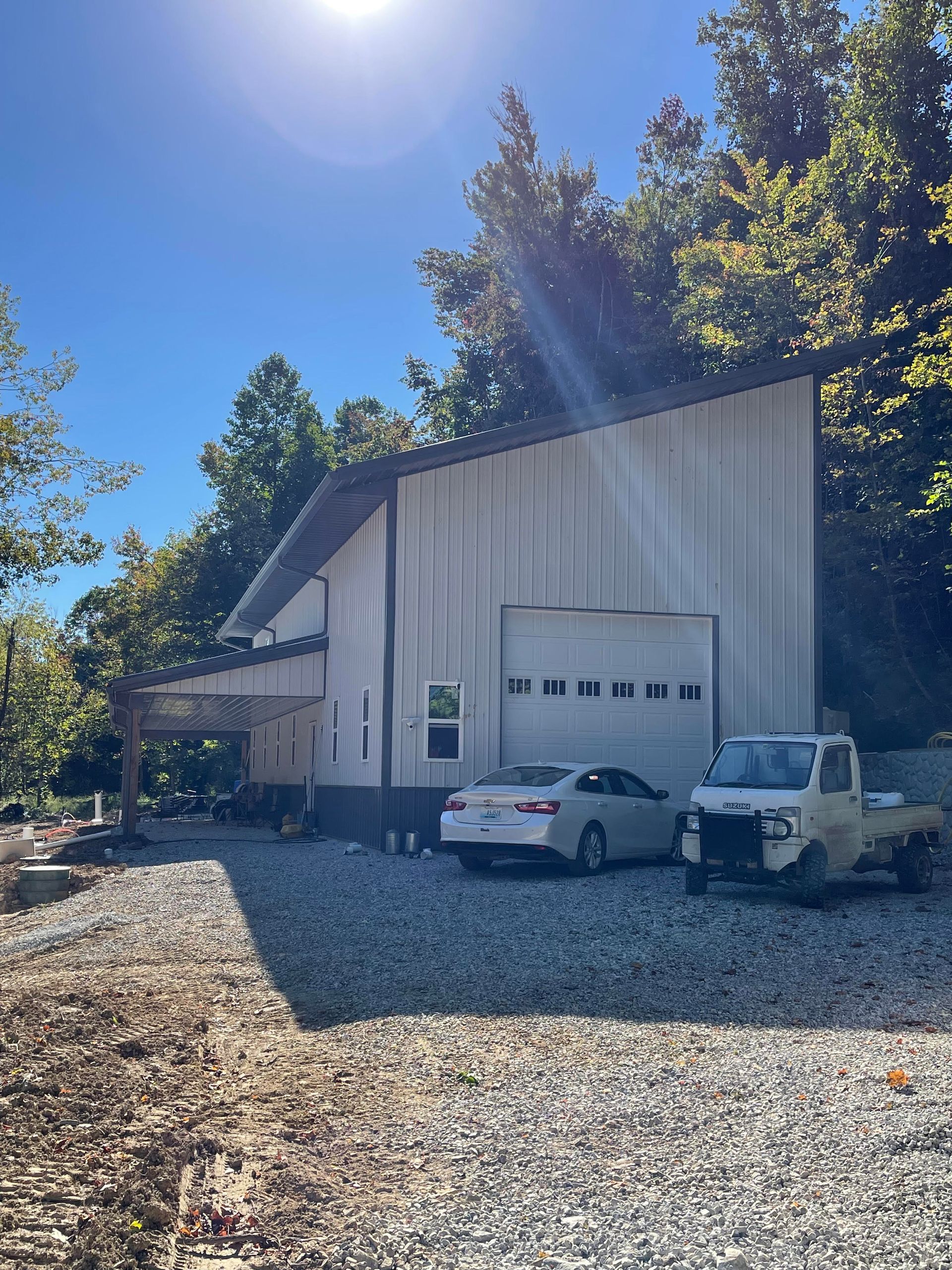 White metal building with a garage door and a covered patio. A car and a small truck are parked in front on a gravel driveway.