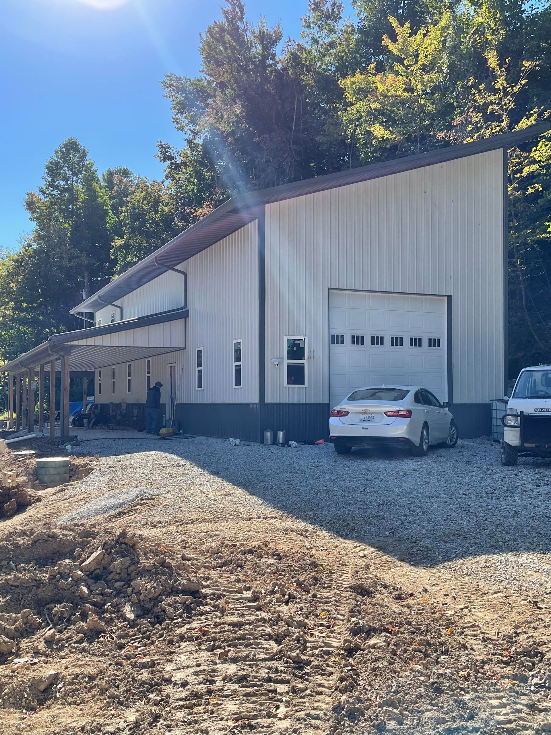 A white metal building with an open garage door, a covered porch, and vehicles parked outside, set against a backdrop of trees under a blue sky.