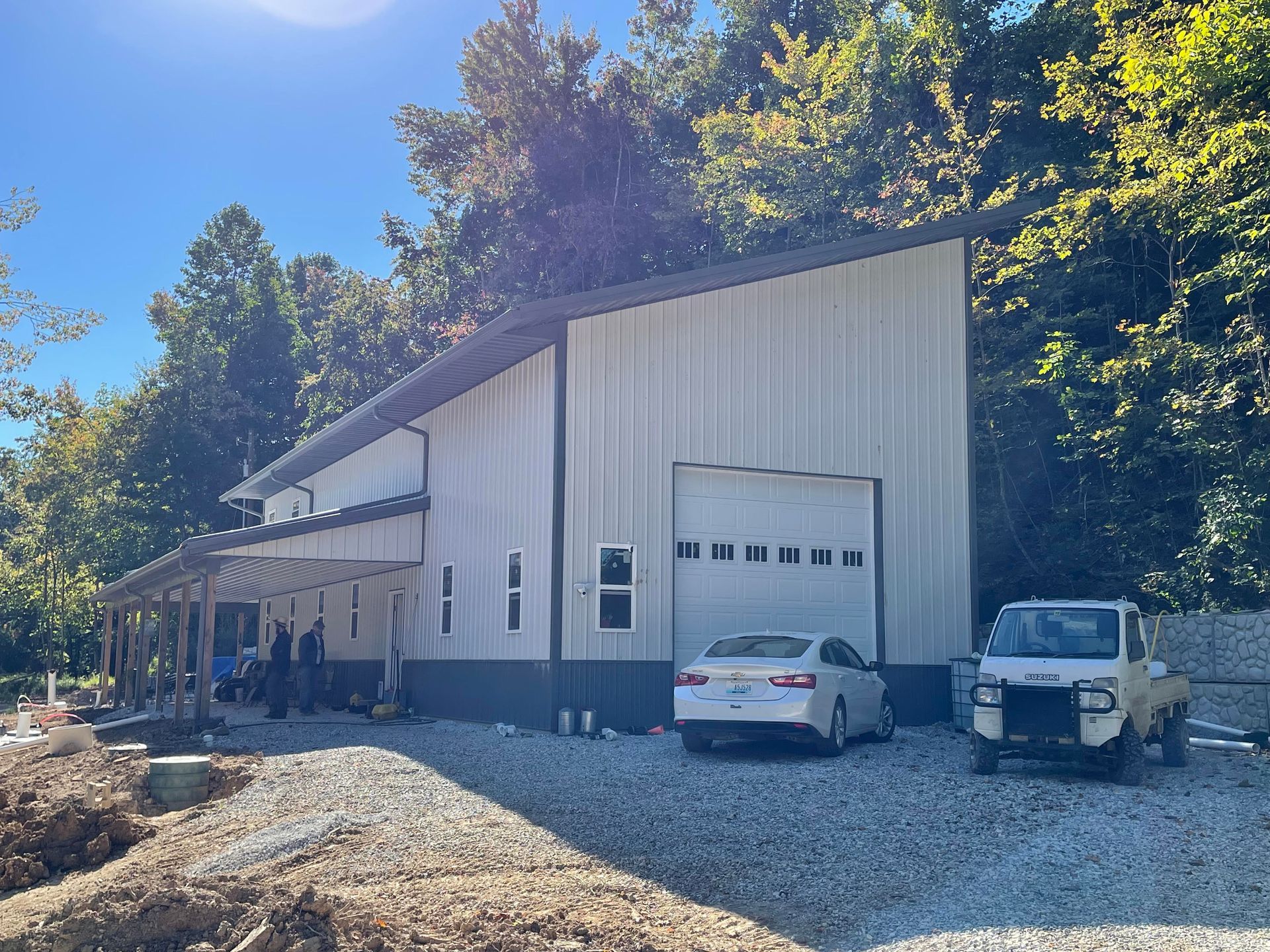 A large metal building with a garage door, a white car, and a white truck parked outside on a gravel driveway.