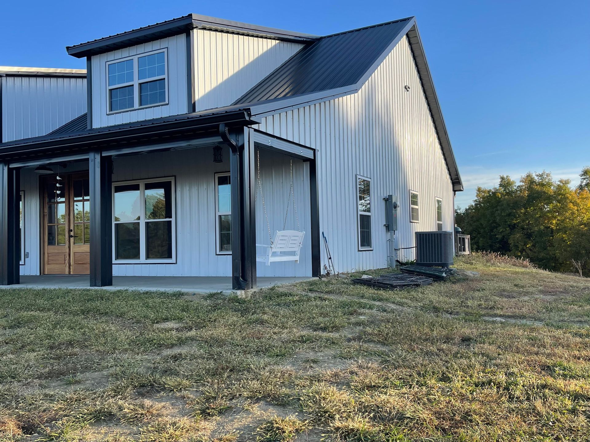 A modern metal-clad house with a black roof and porch, set in a grassy field against a blue sky.