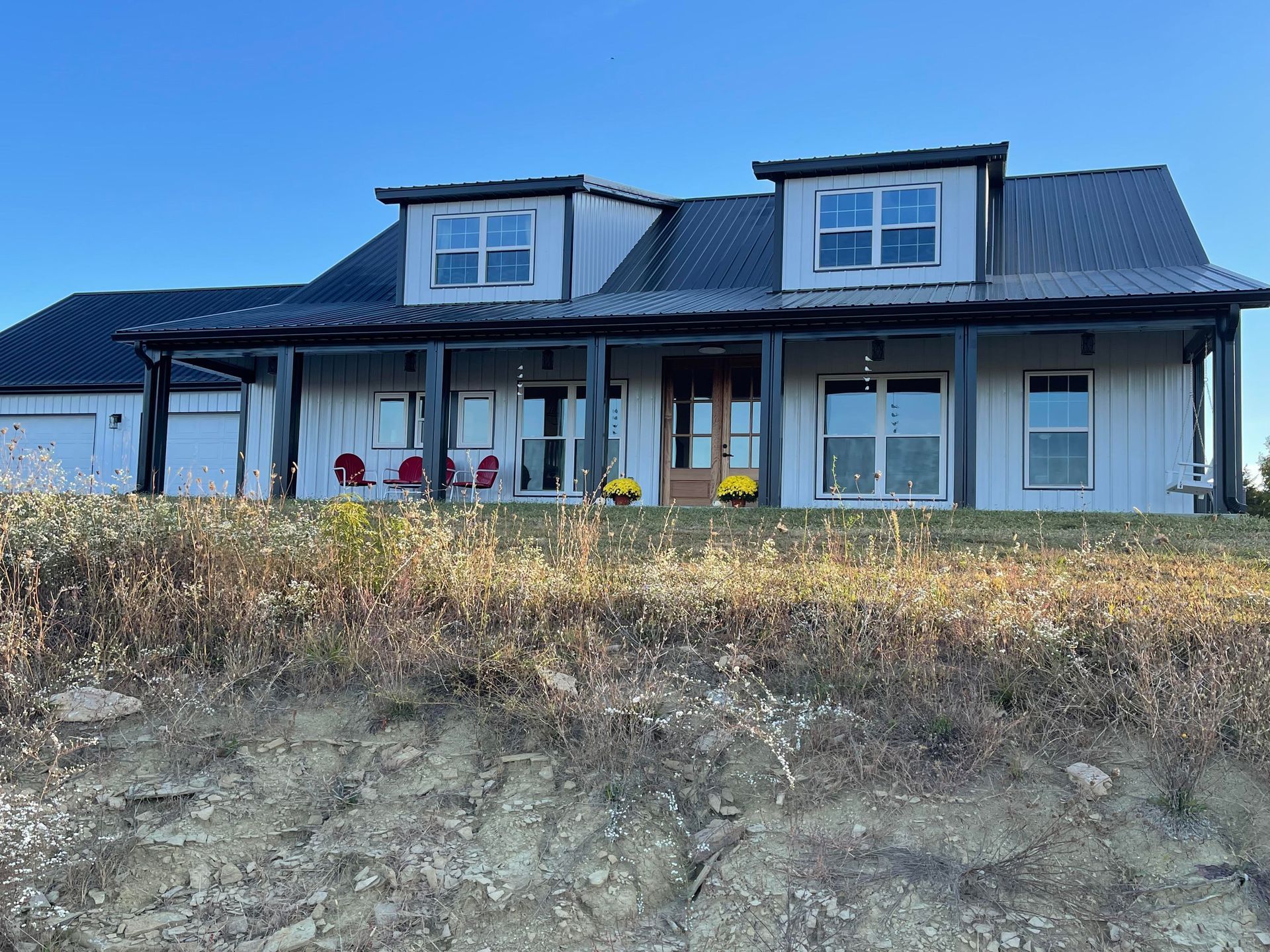 A light gray farmhouse with a dark roof and black trim, with a porch and dormer windows, set against a blue sky.