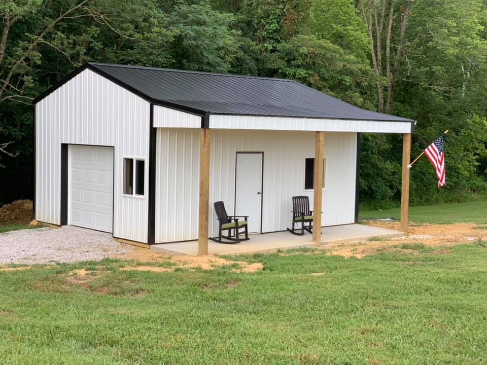 White metal building with black roof, porch, garage door, and American flag. Two rocking chairs sit on the porch.